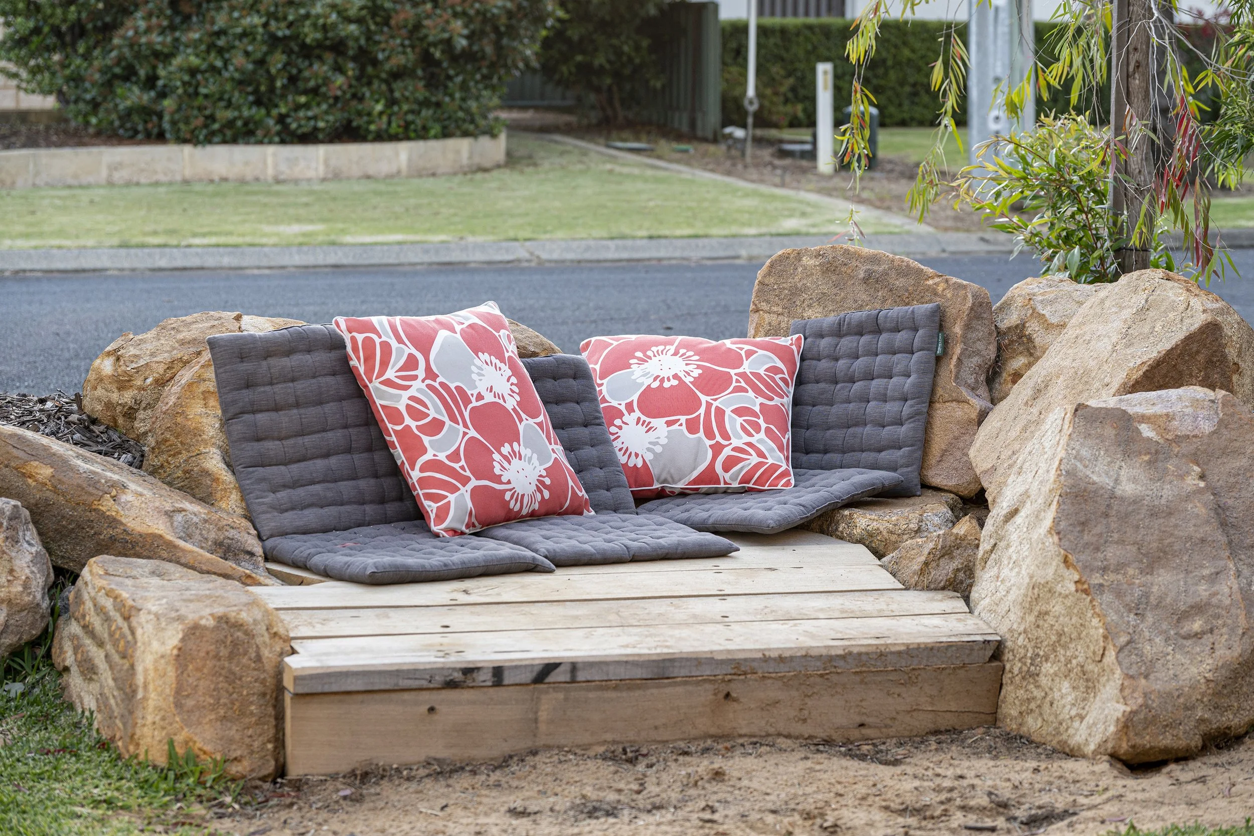 Outdoor seating area with a gray cushioned bench, two orange and white floral pillows, surrounded by large rocks, with a grassy lawn and trees in the background.