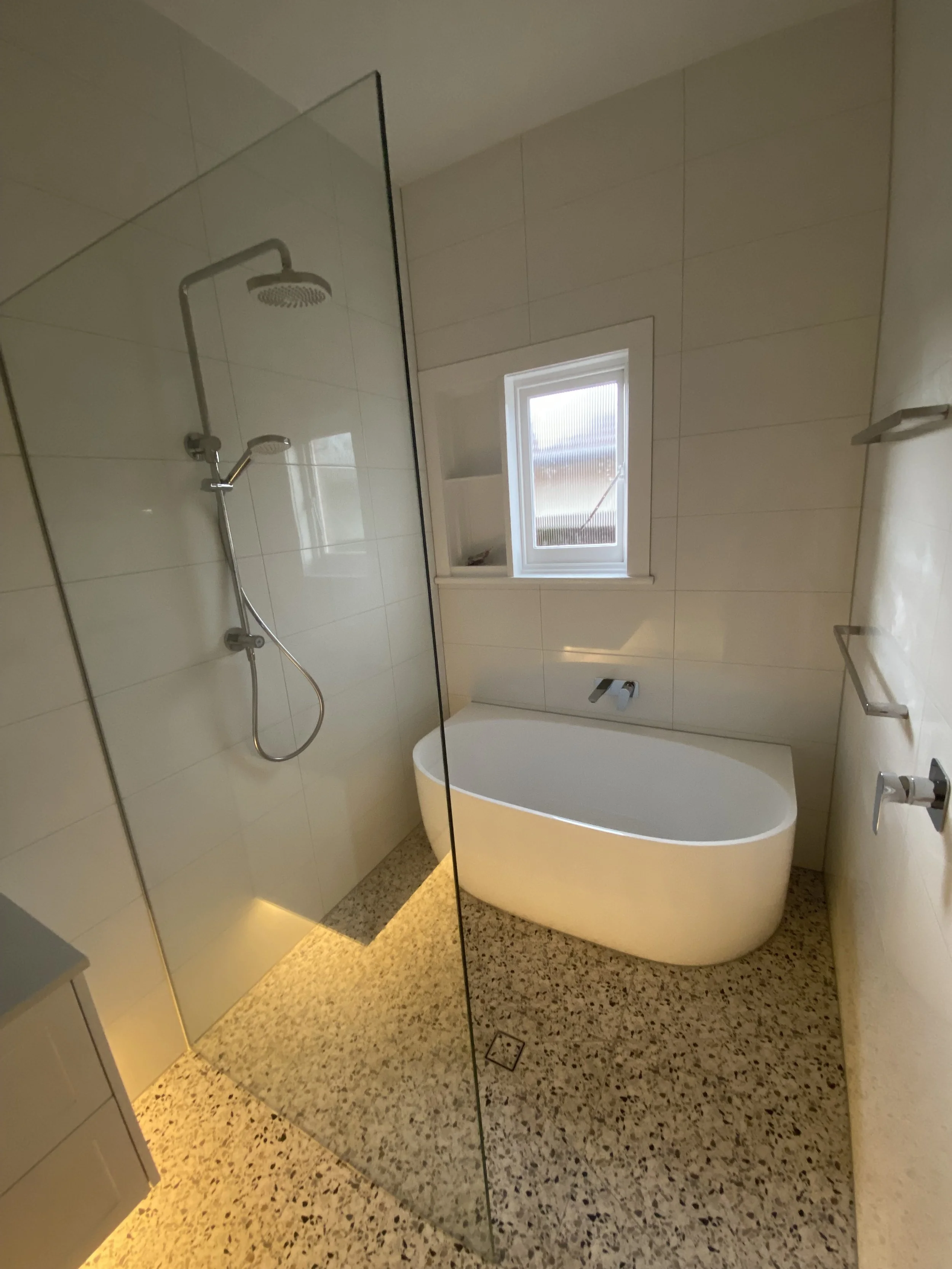 Modern bathroom with a bathtub, a shower, a window, and a speckled terrazzo floor, bathroom fixtures, and white tiled walls.