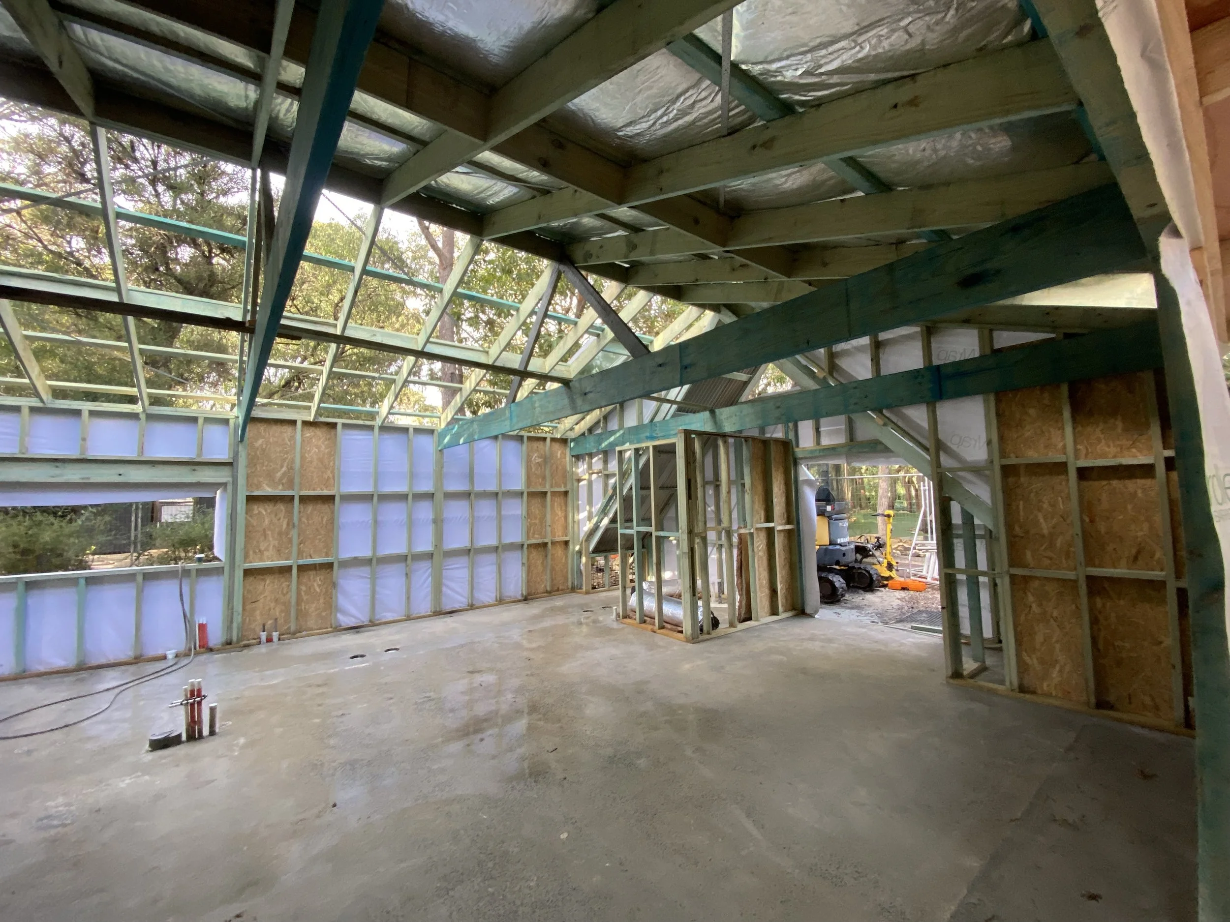 The image shows a house under construction with a wooden frame and an unfinished roof. The interior is open with visible studs and beams, and construction tools and materials are on the floor.