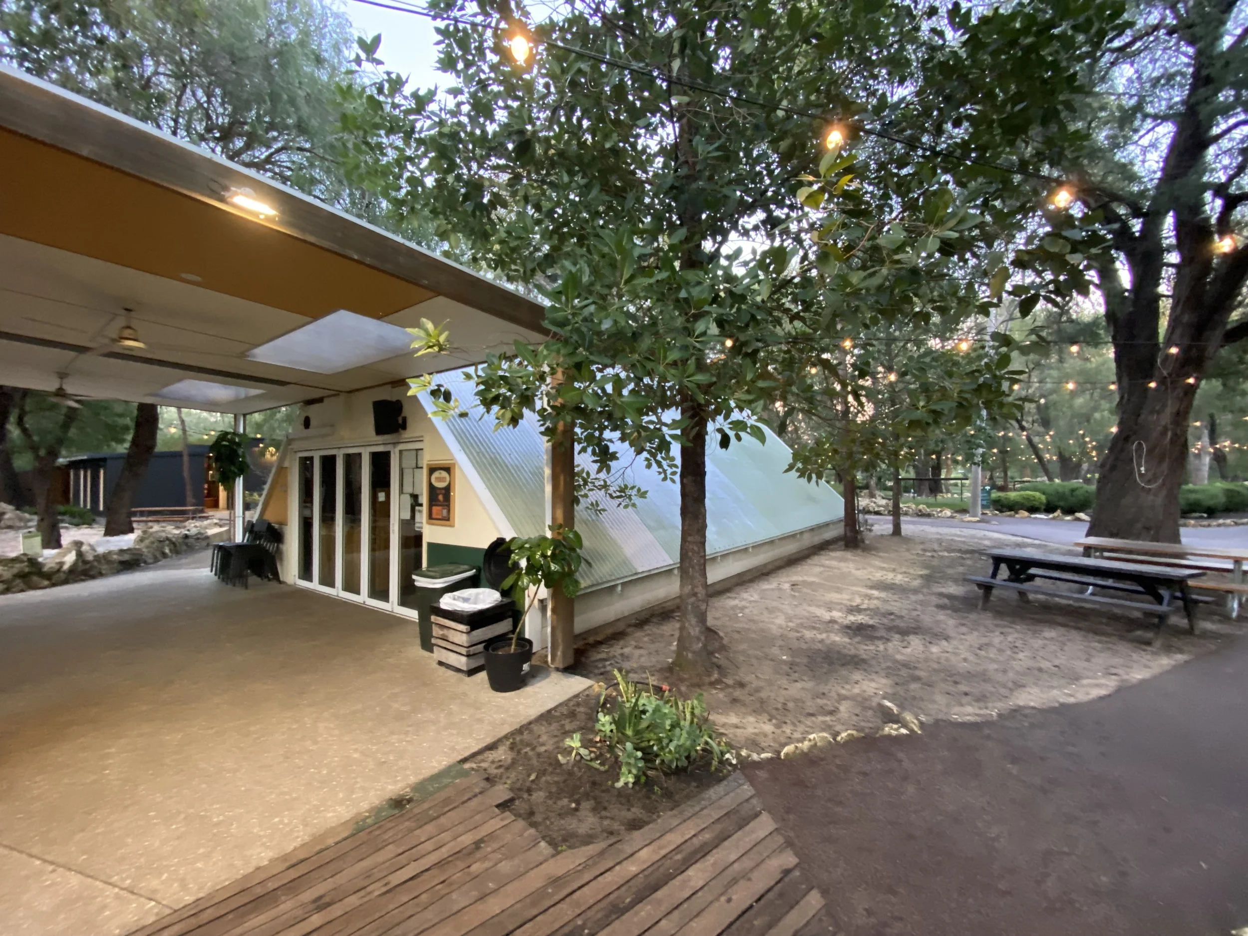An outdoor scene during sunset with string lights hanging above. A small building or structure with a door and windows, surrounded by trees and benches. Some plants and a trash bin are also visible.