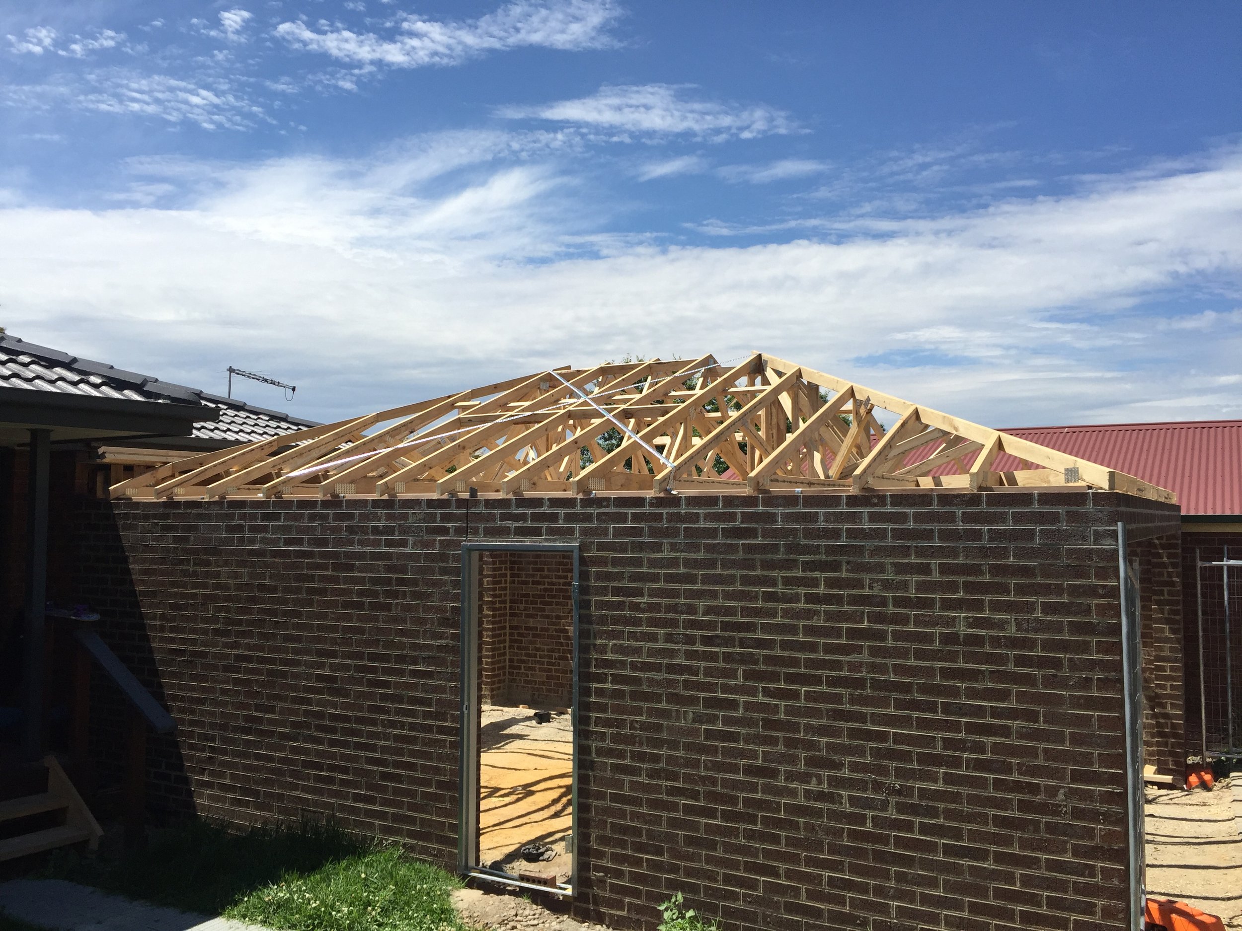 Construction site with a brick wall and wooden roof framing under a blue sky with clouds.