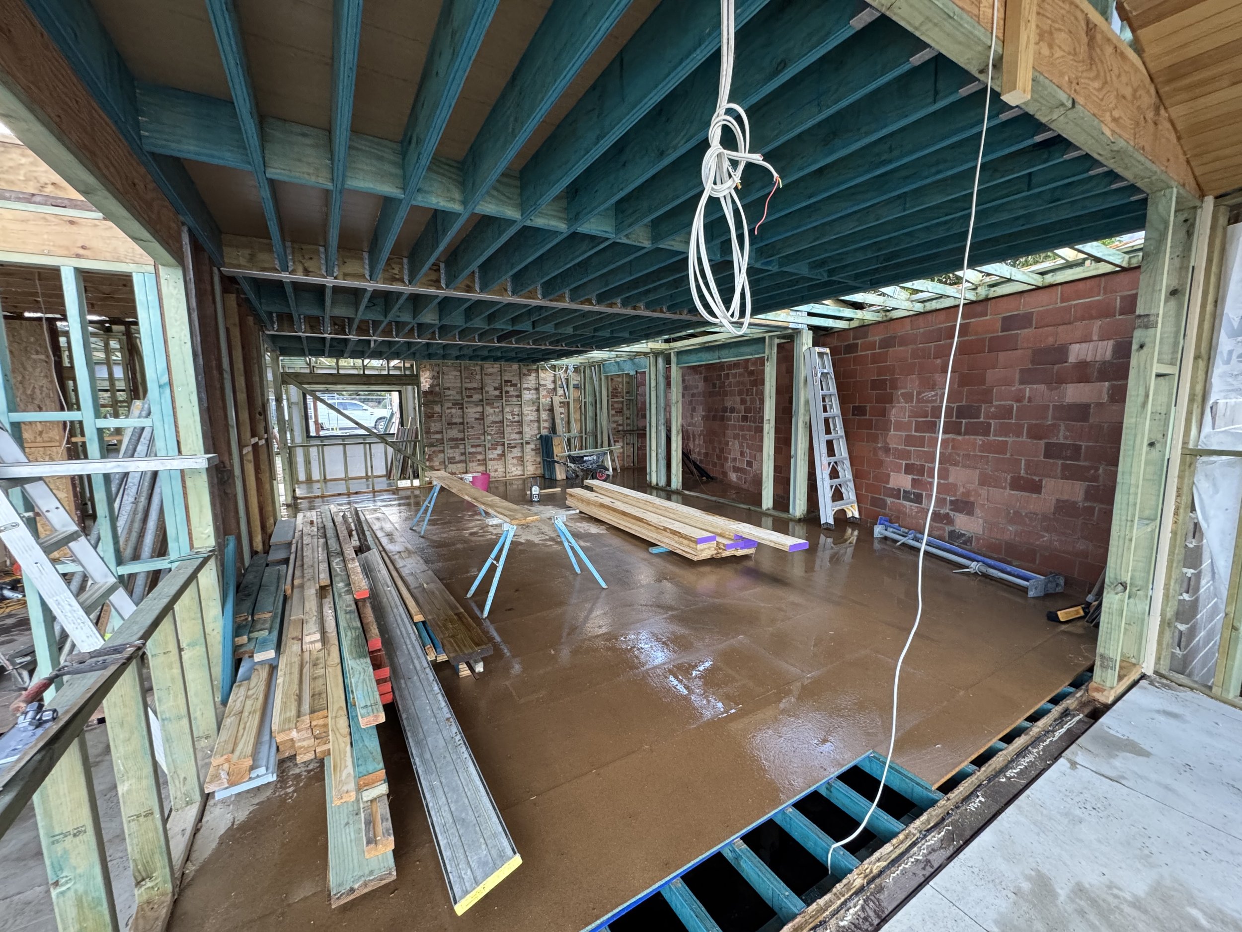 Construction site with wooden framework and brick wall, electrical wiring hanging from ceiling, and tools and lumber on the floor.