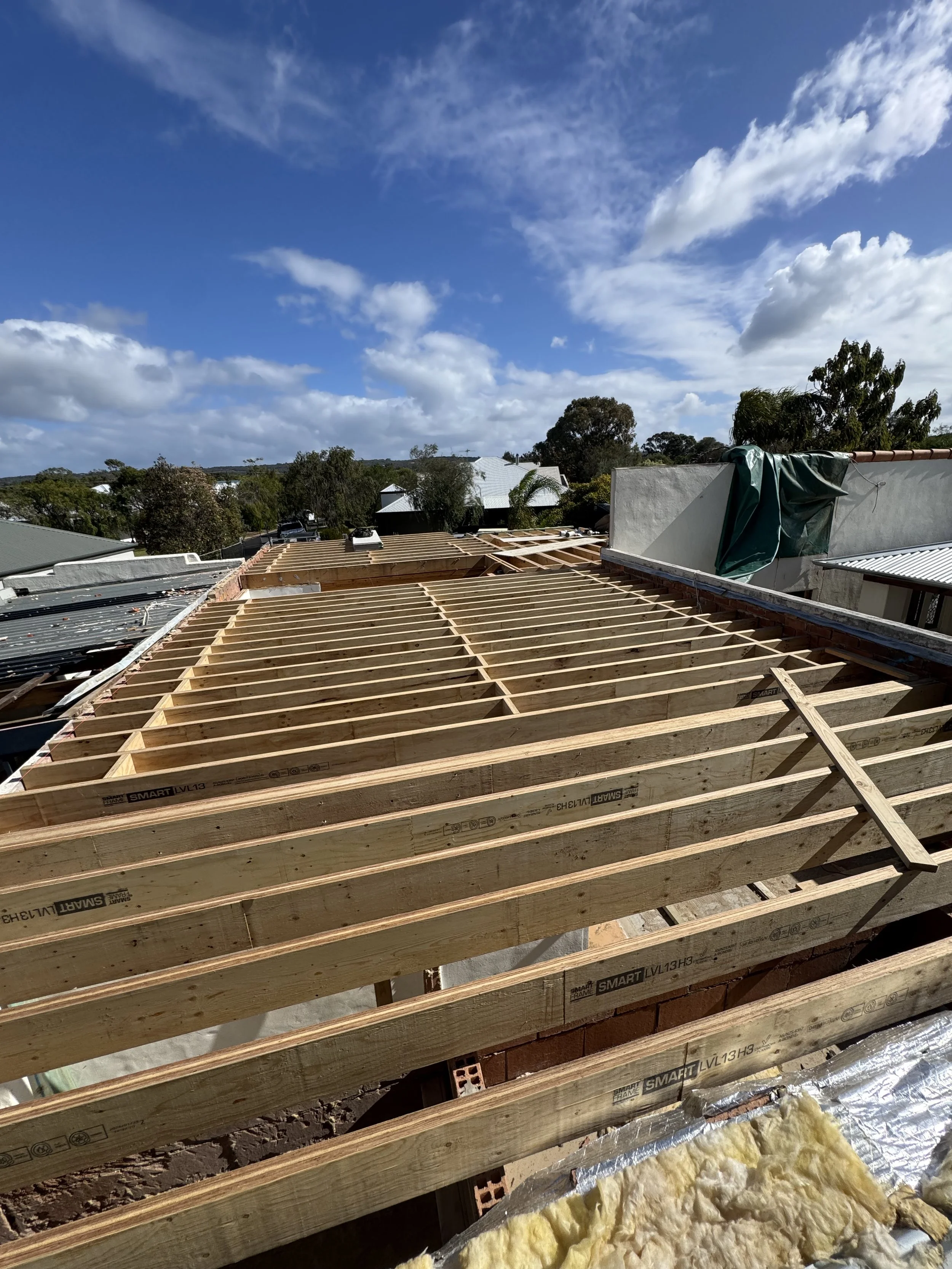 Construction site with wooden roof framework, insulation material below, and houses with trees and a blue sky with clouds in the background.