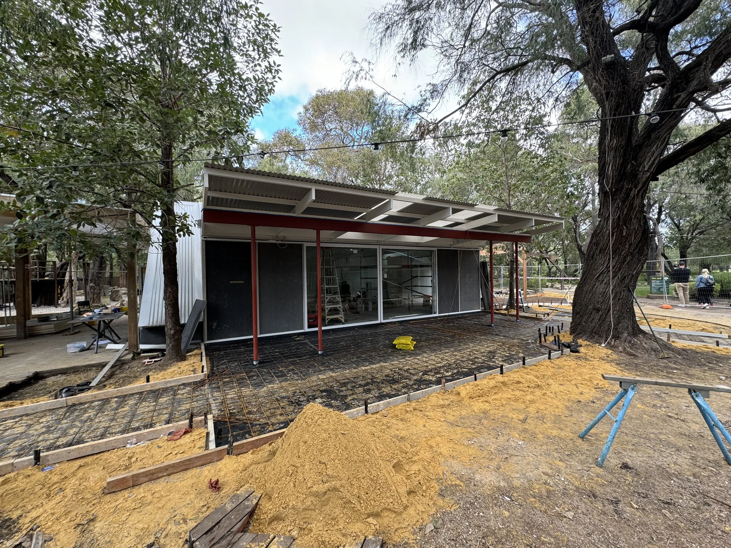 Construction site with building frame, stacks of sand, and ground prepared for concrete, surrounded by trees.