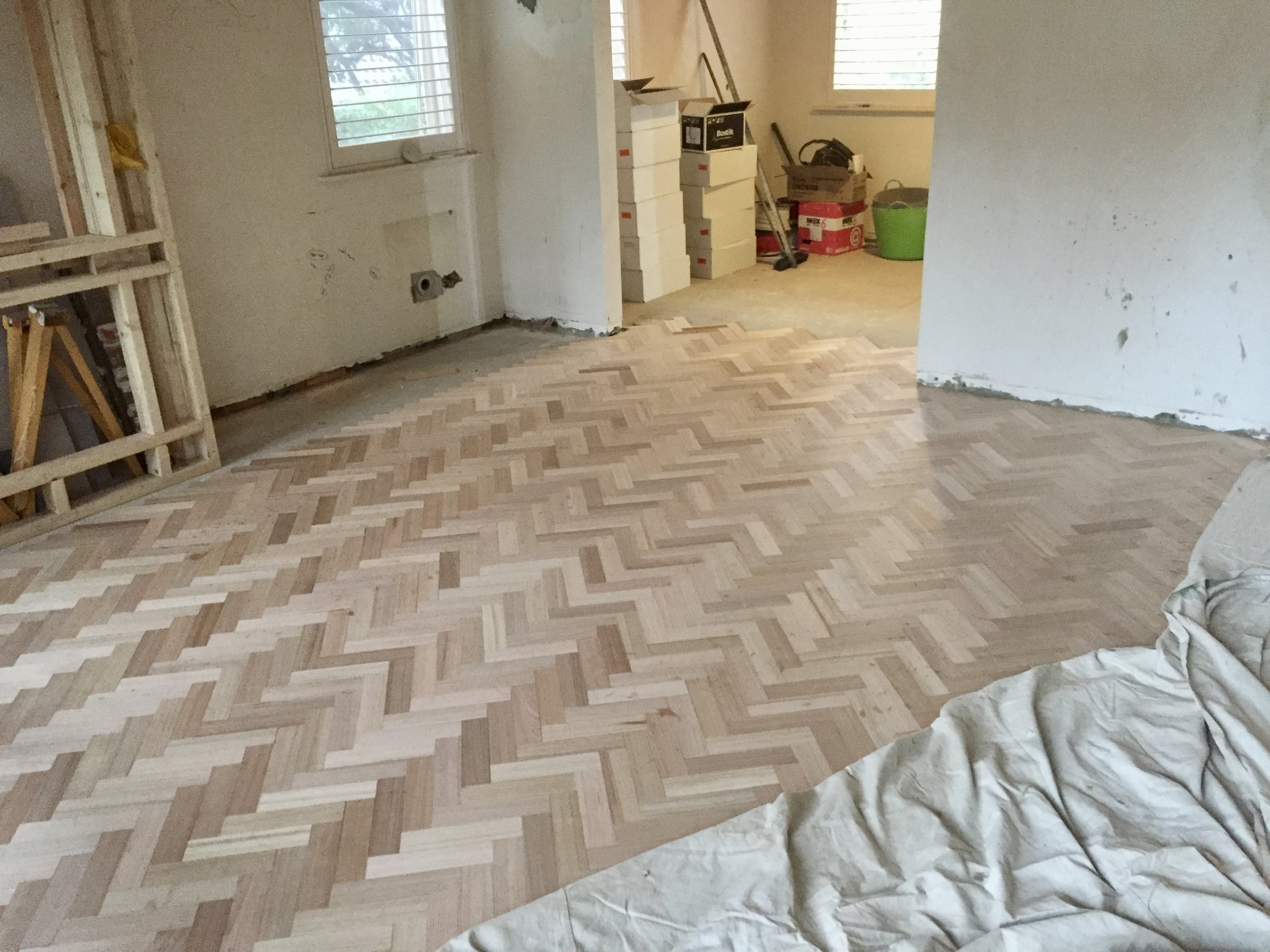 An unfinished room with a new wooden herringbone floor. Construction materials and tools are scattered around, including stacks of white boxes, a green bucket, and a ladder.