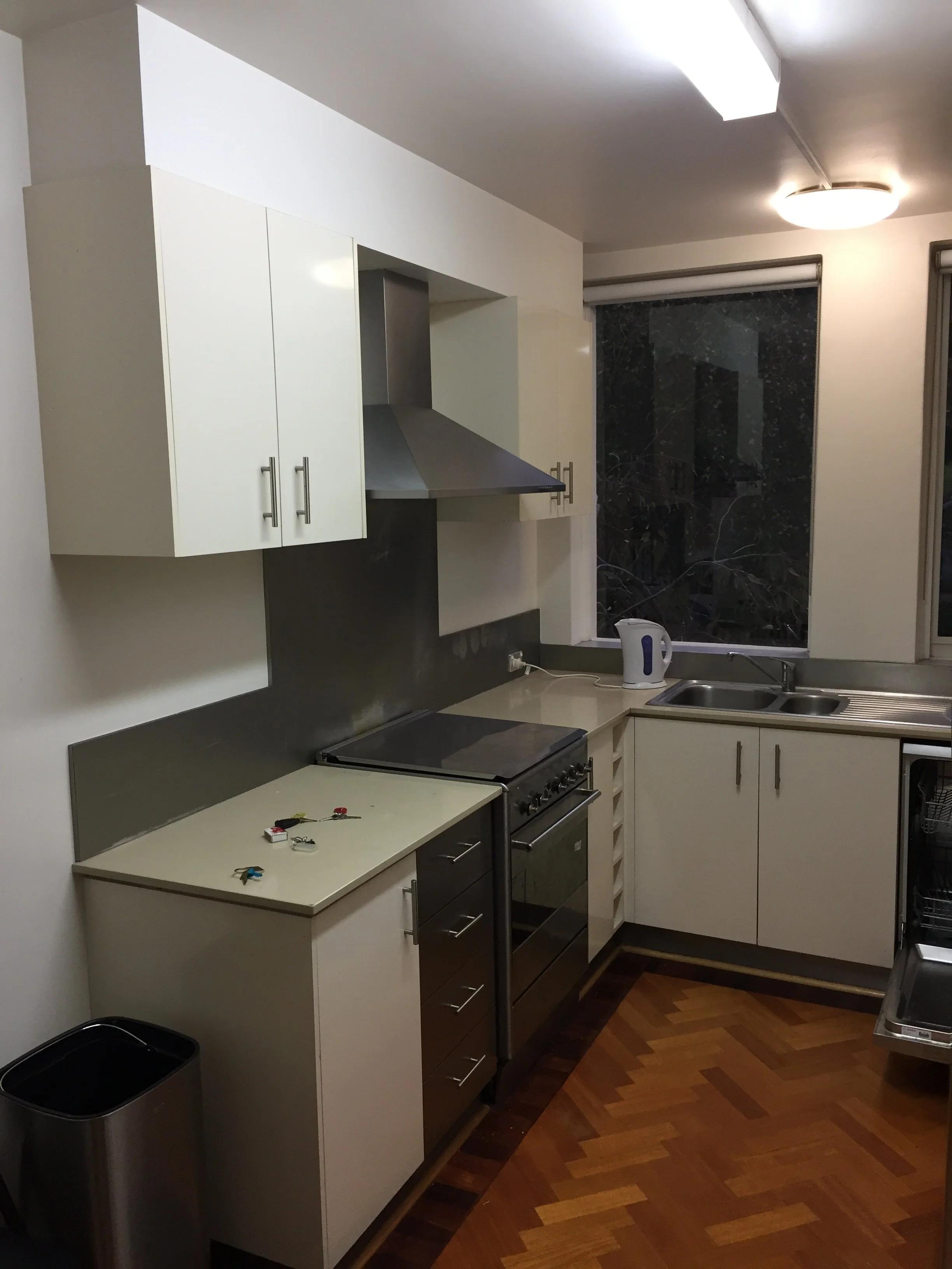 A small kitchen with white cabinets, a stove, a sink, and a window with a dark marble curtain. There is a white electric kettle on the countertop and some keys and small items on the counter. The floor has wooden parquet.