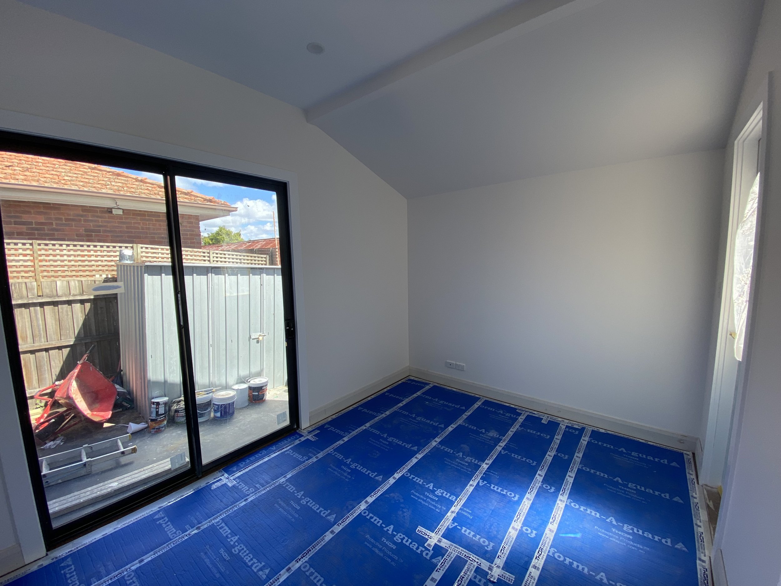An unfinished room with a large sliding glass door leading outside. The room has white walls and a blue protective floor covering with labels. Outside, there is a backyard enclosed by a wooden fence, with construction tools and materials visible near