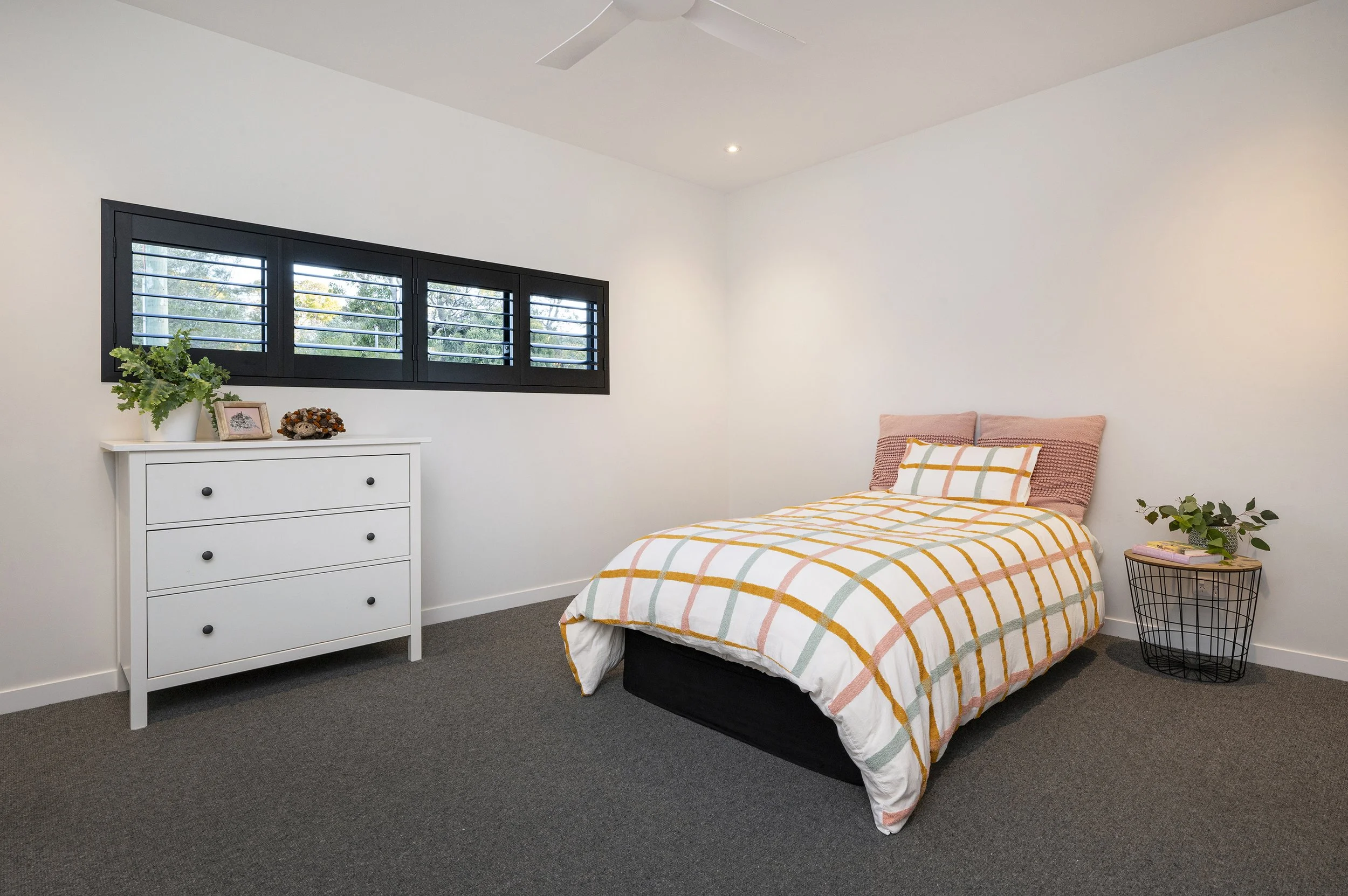 Bedroom with white walls, a window with black shutters, a white dresser with decorative items, a bed with a yellow, orange, and green plaid bedspread, pink pillows, and a side table with plants and books.