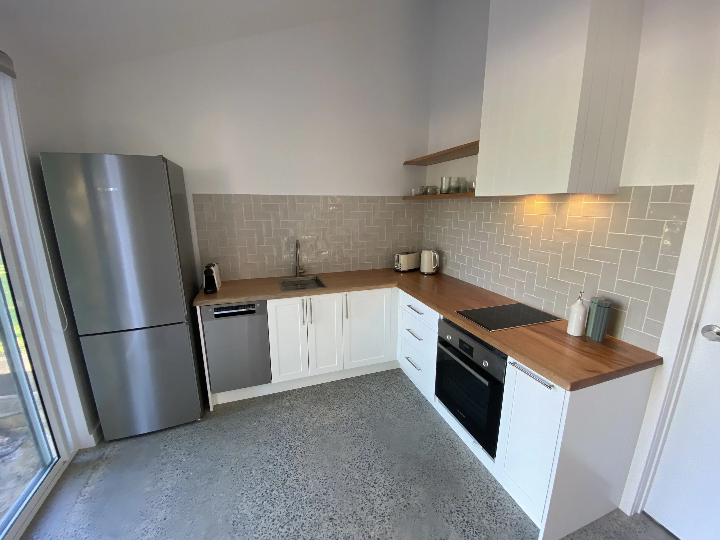 Modern kitchen with white cabinets, wooden countertop, gray tile backsplash, stainless steel refrigerator, toaster, kettle, oven, and large windows.