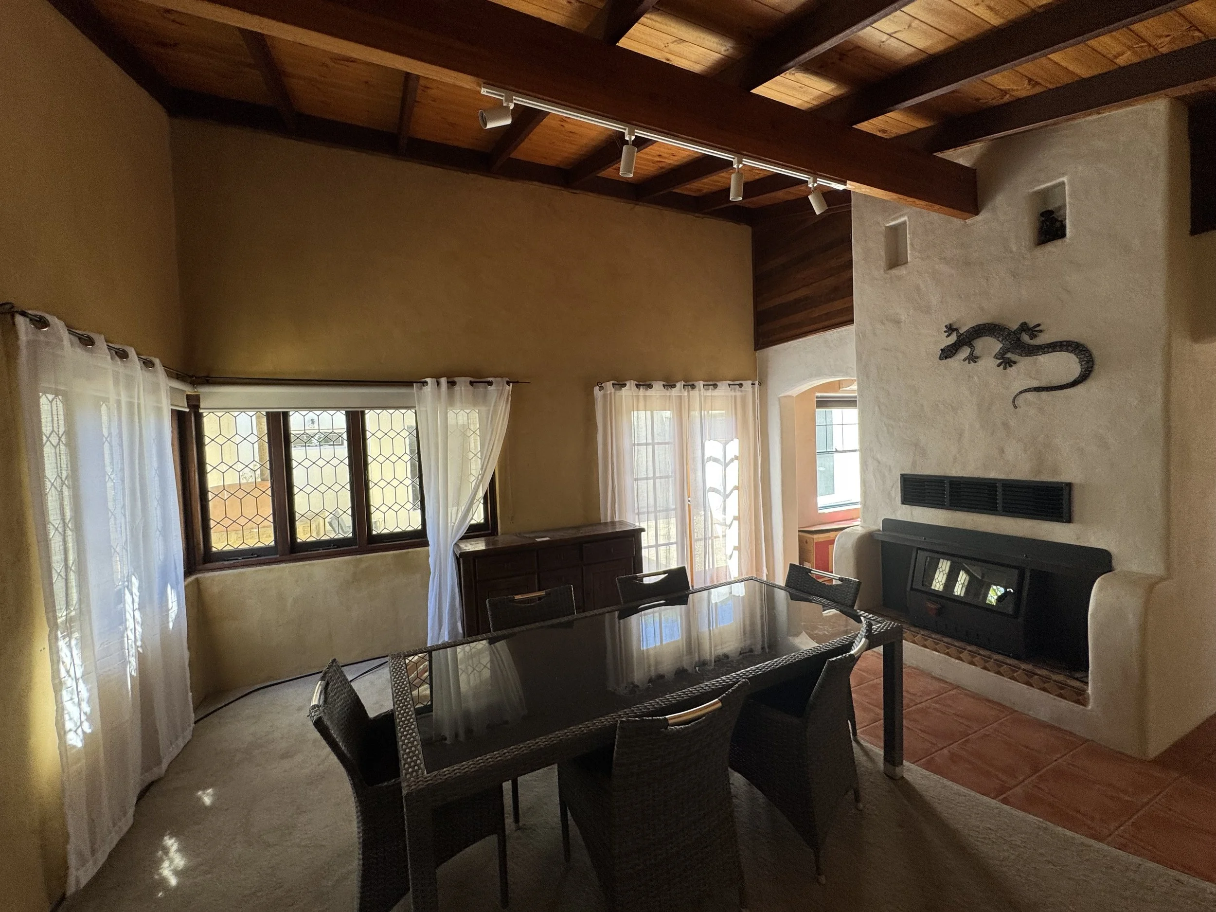 A living room with beige-colored walls, large windows with white curtains, a wooden dining table with dark woven chairs, a fireplace, a wall-mounted lizard decoration, and a ceiling with exposed wooden beams and track lighting.
