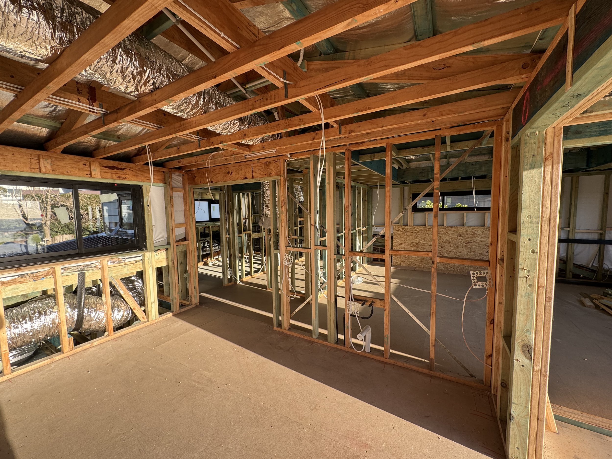 Interior view of a house under construction showing exposed wooden framing, ductwork, electrical wiring, and large windows.