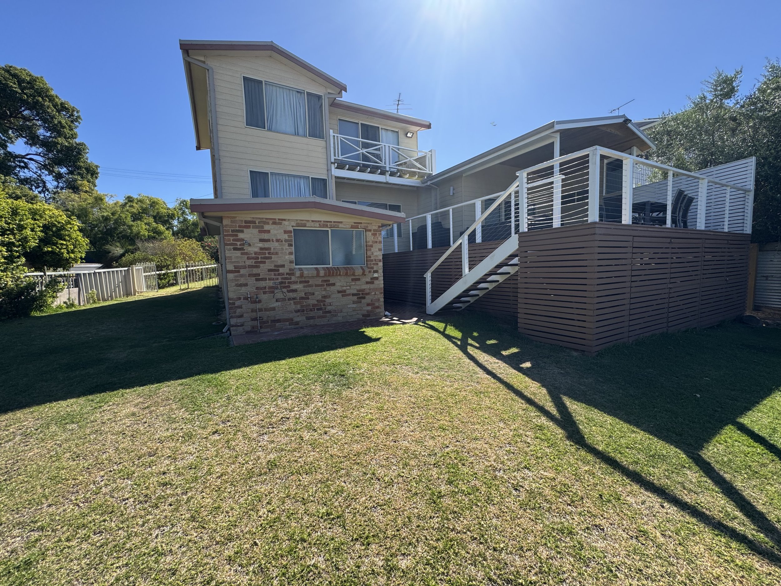 A house with a brick foundation and cream-colored siding, featuring large windows, a white balcony, and a wooden deck with outdoor furniture, surrounded by green trees and a fenced yard under a clear blue sky.