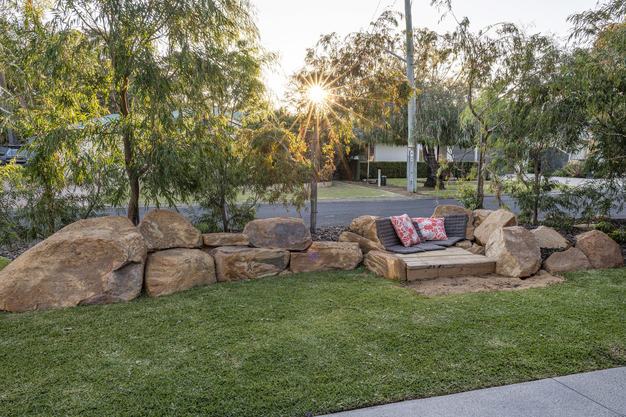 A backyard during sunset with a stone retaining wall, a cushioned outdoor seating area with decorative pillows, lush trees, and a manicured lawn.