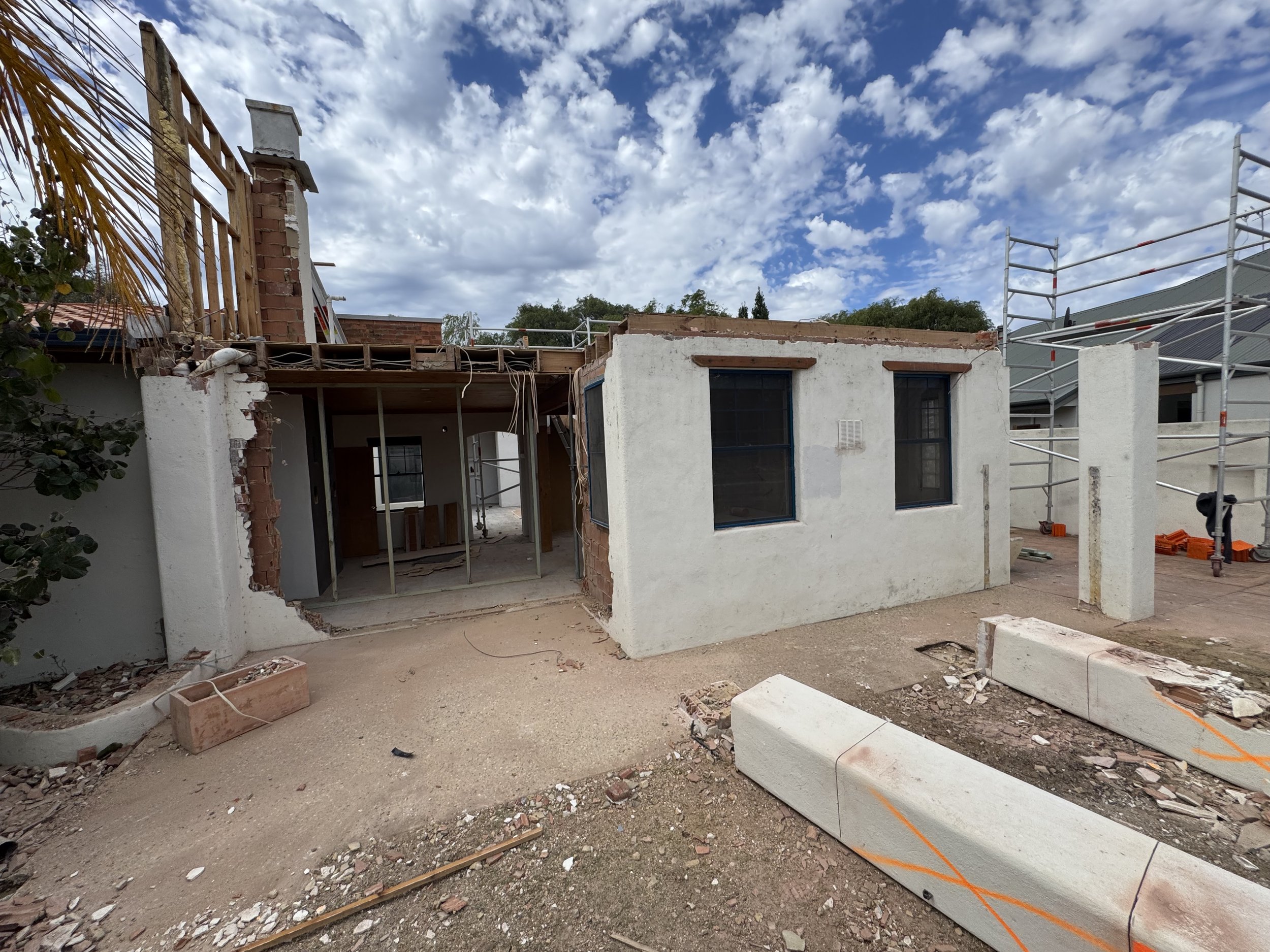 House under construction with scaffolding, partially built walls, and construction materials on the ground, under a partly cloudy sky.