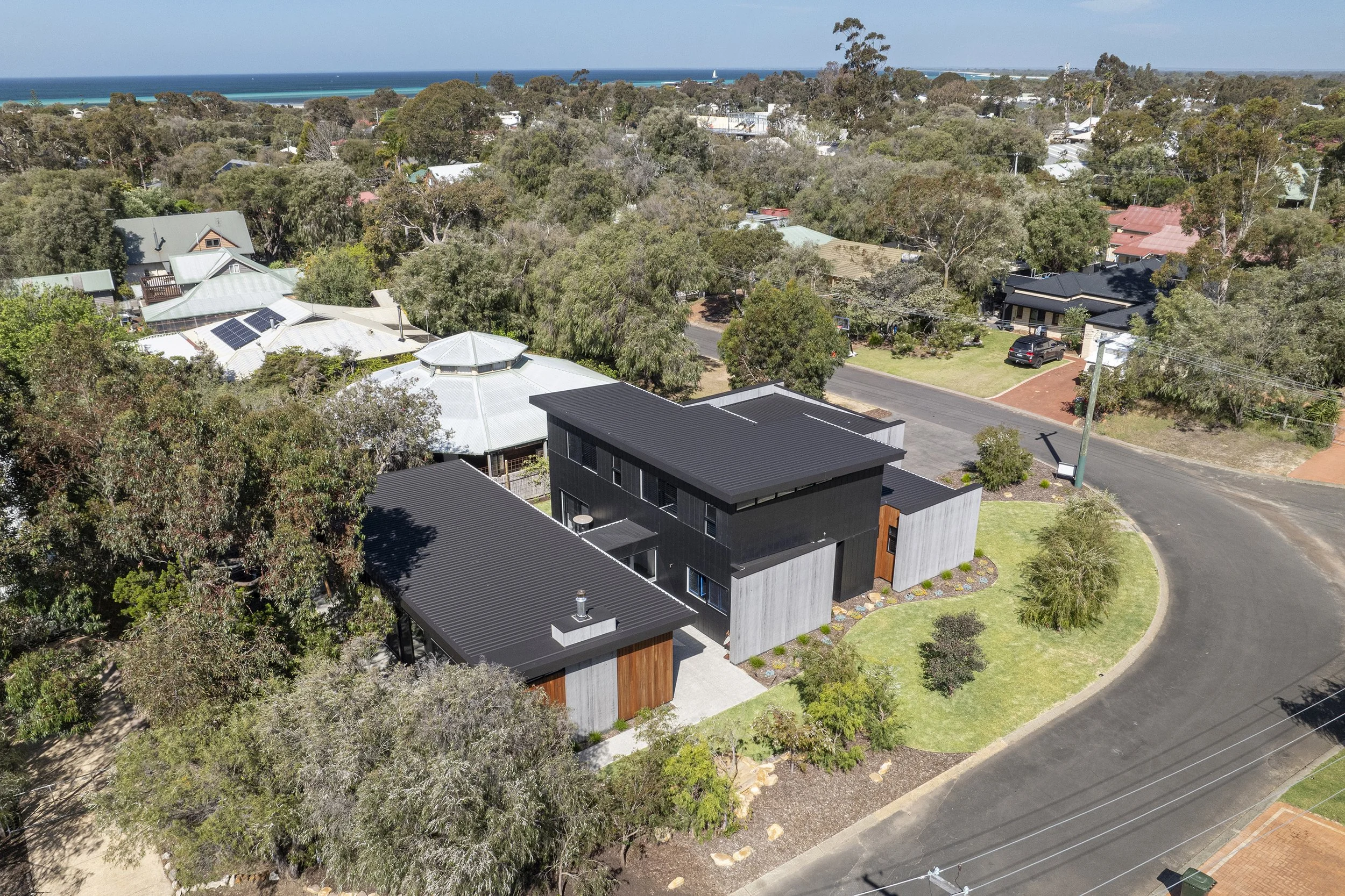Aerial view of a residential neighborhood with a mix of modern and traditional houses, surrounded by trees and greenery, near the coastline with visible ocean and sailboats in the distance.