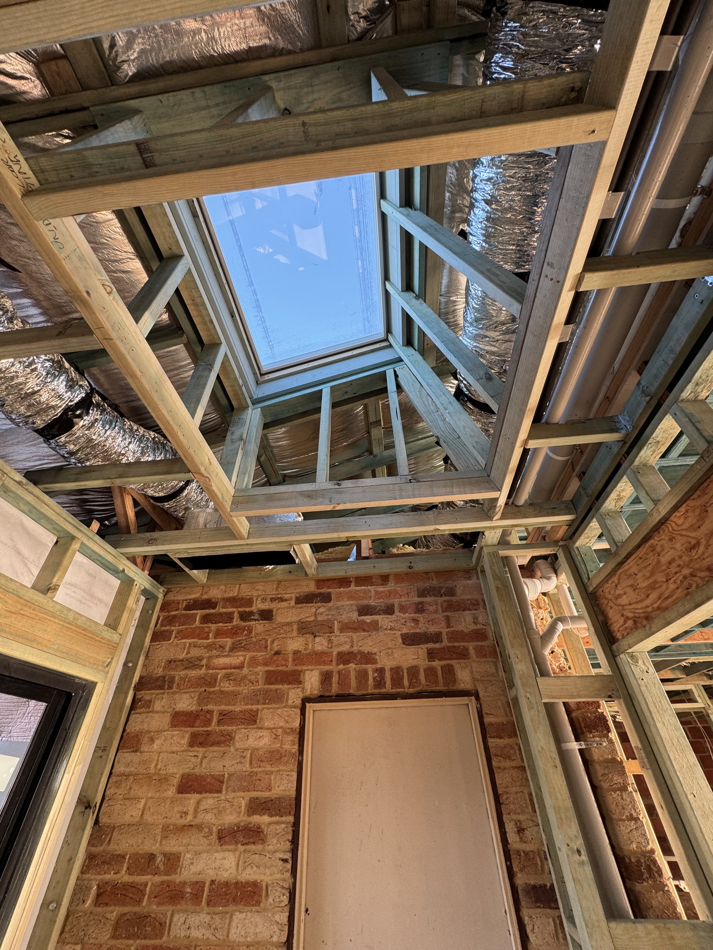 Interior view of a building under construction, showing wooden framework, metal ducting, a large rectangular window, and a brick wall.