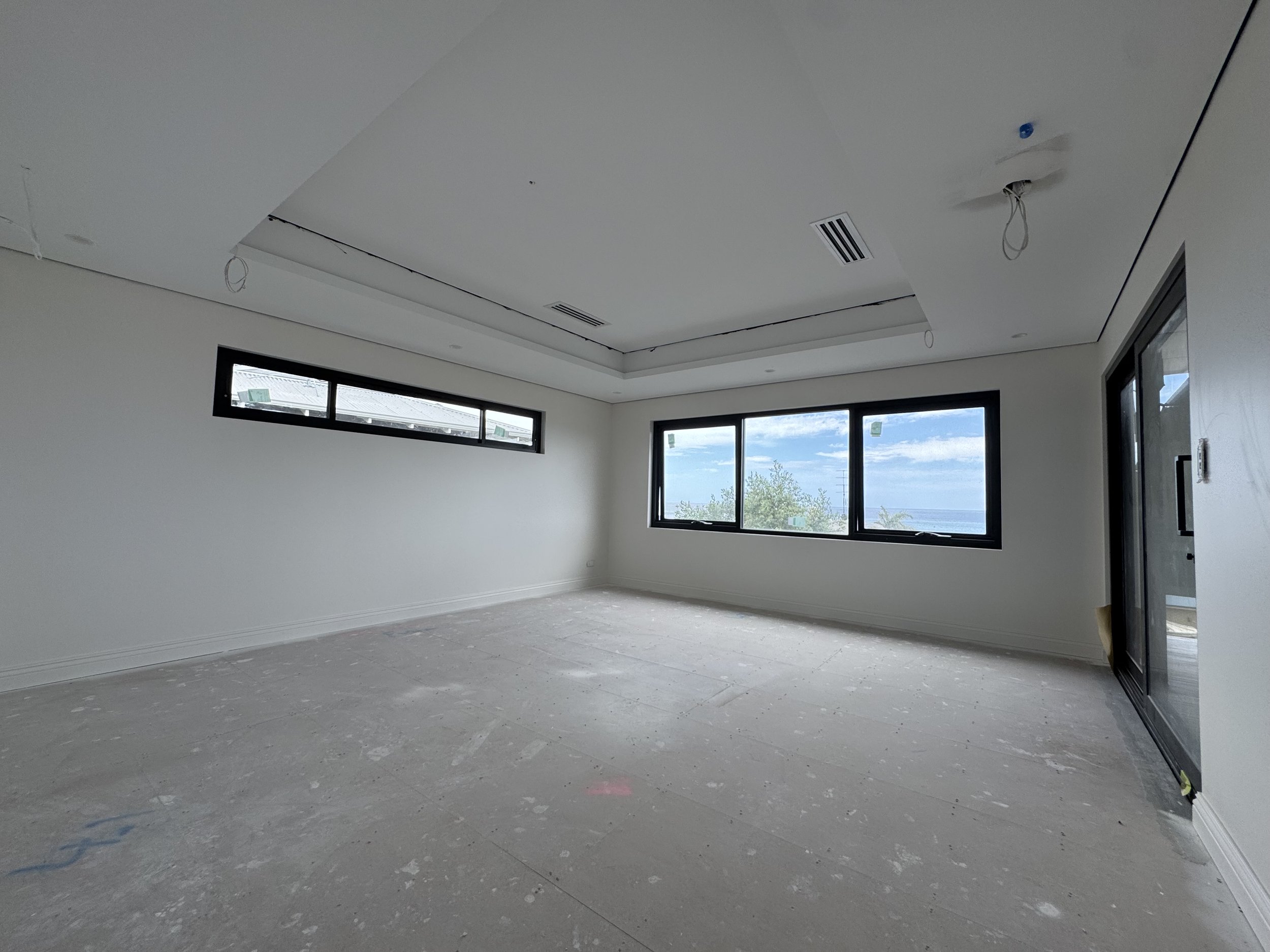 Empty room under construction with unfinished flooring, white walls, large windows, and hanging wires from the ceiling.