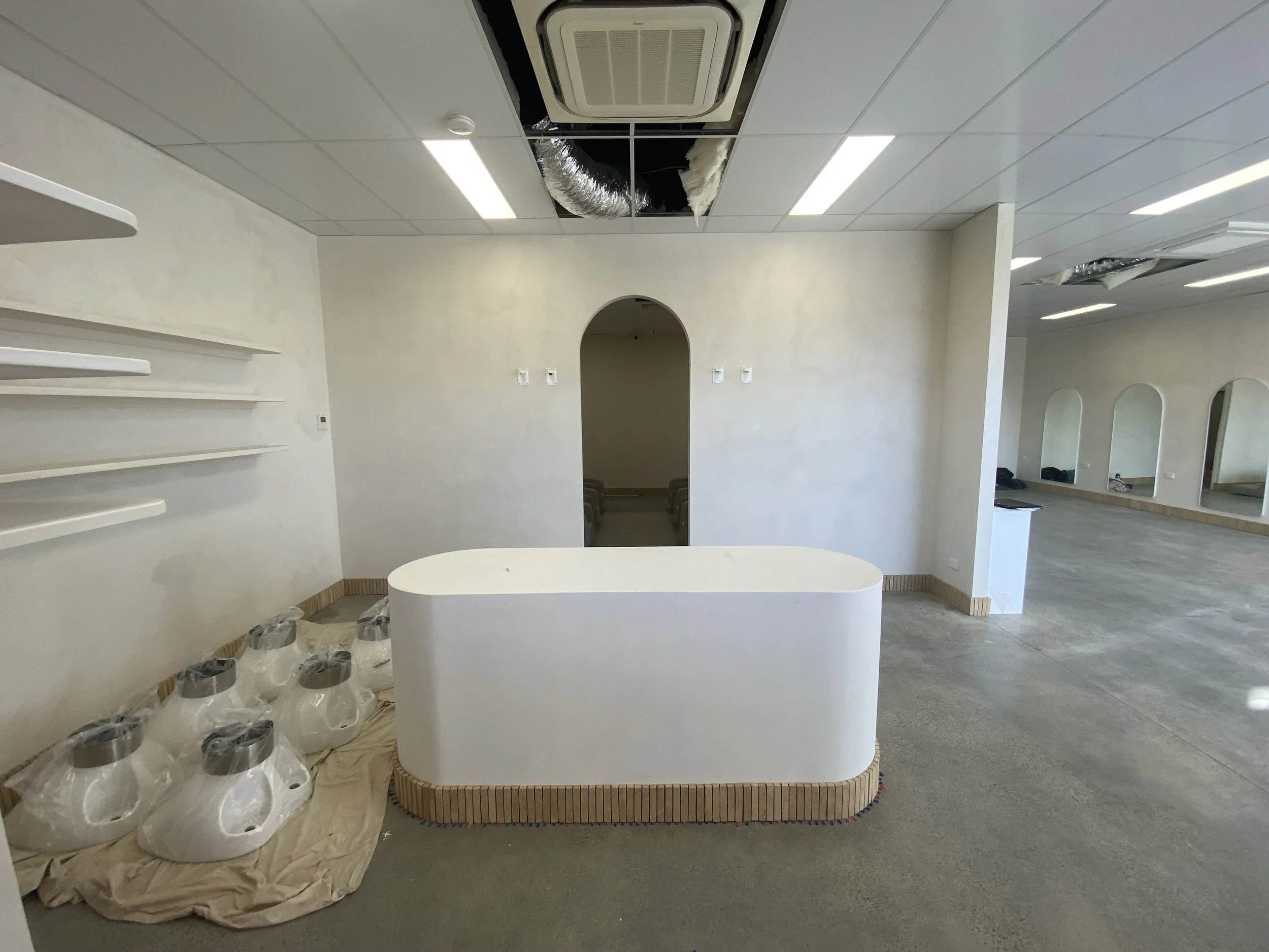 Room under renovation with white walls, new ceiling panels, ceiling air conditioning unit, and open ceiling tiles exposing ductwork and insulation. Several plastic containers are covered with paper and lay on the floor.