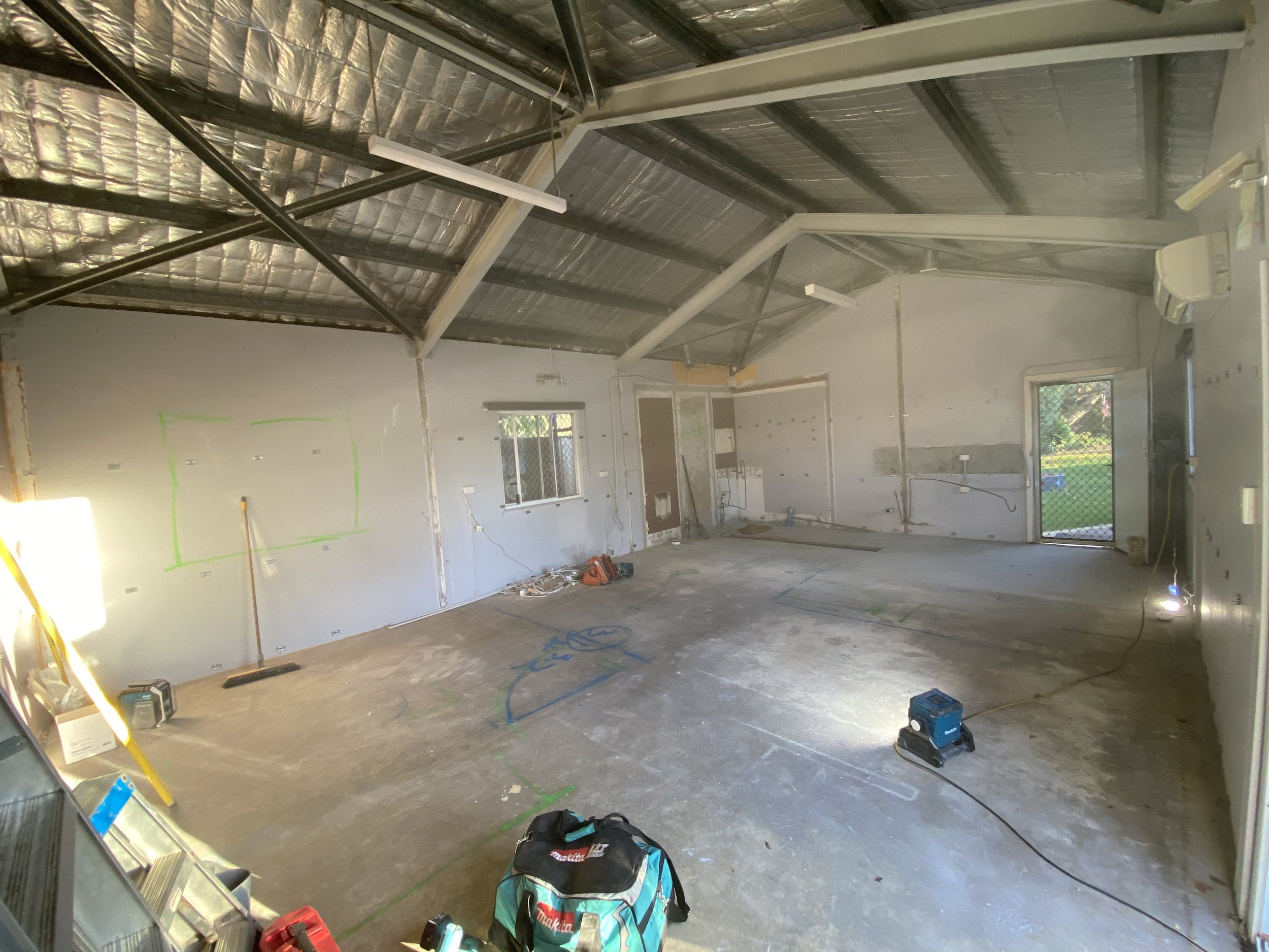 Construction site inside a building with exposed ceiling framing, insulation, and unfinished walls. Tools and construction materials are scattered on the floor.