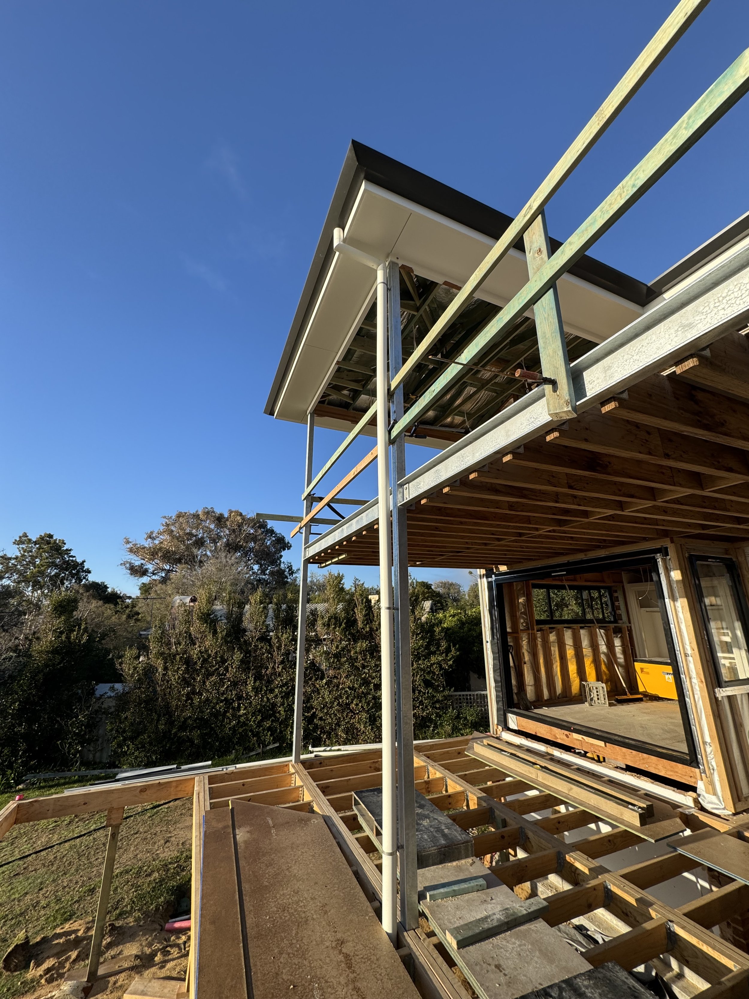 Construction site with a partially built second-story deck and scaffolding, showing the underside of a roof and framing, with trees and a clear blue sky in the background.