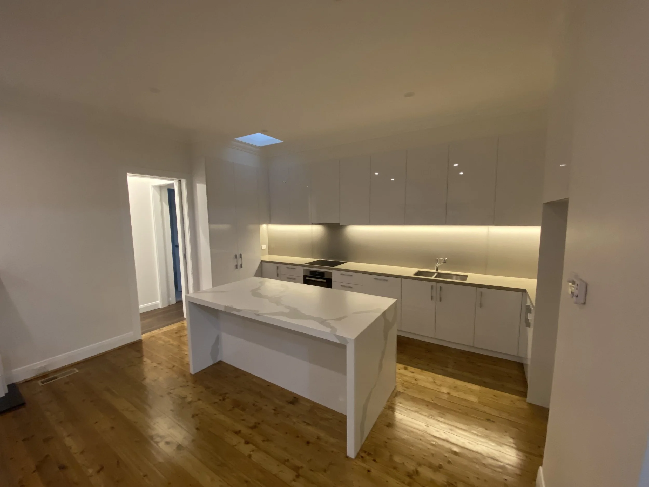 A modern kitchen with white cabinets, a kitchen island, hardwood floors, a small rectangular window, and a view into another room.