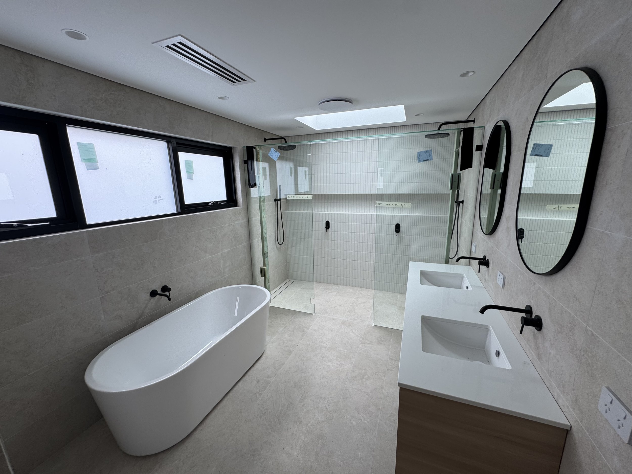 A modern bathroom with a freestanding bathtub, two oval mirrors, a sink with a black faucet, a glass shower enclosure, and a skylight. There are windows on one wall.