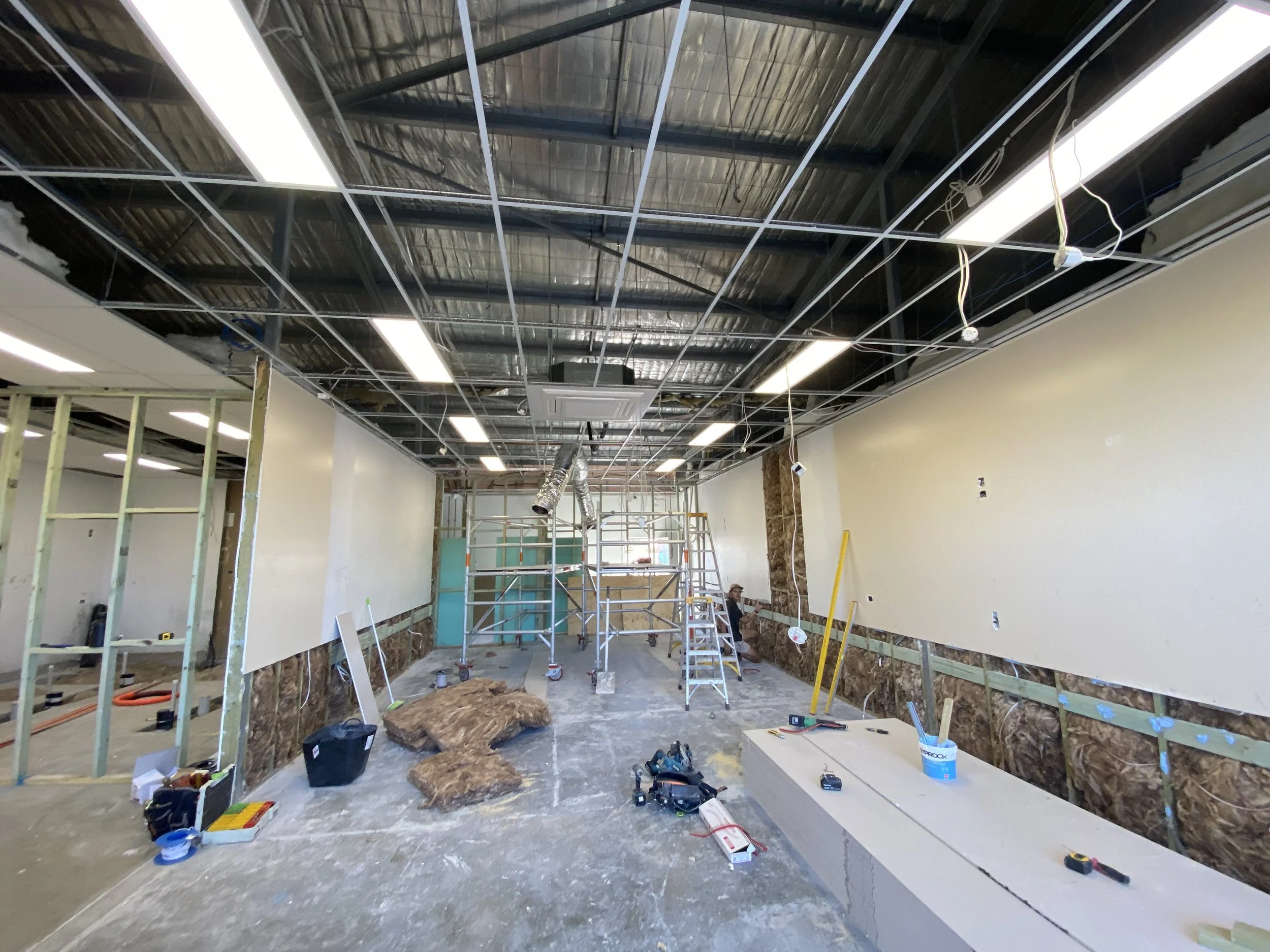 Construction site interior showing an unfinished ceiling with metal framing, bright fluorescent lights, exposed wiring, and construction tools scattered on the floor.