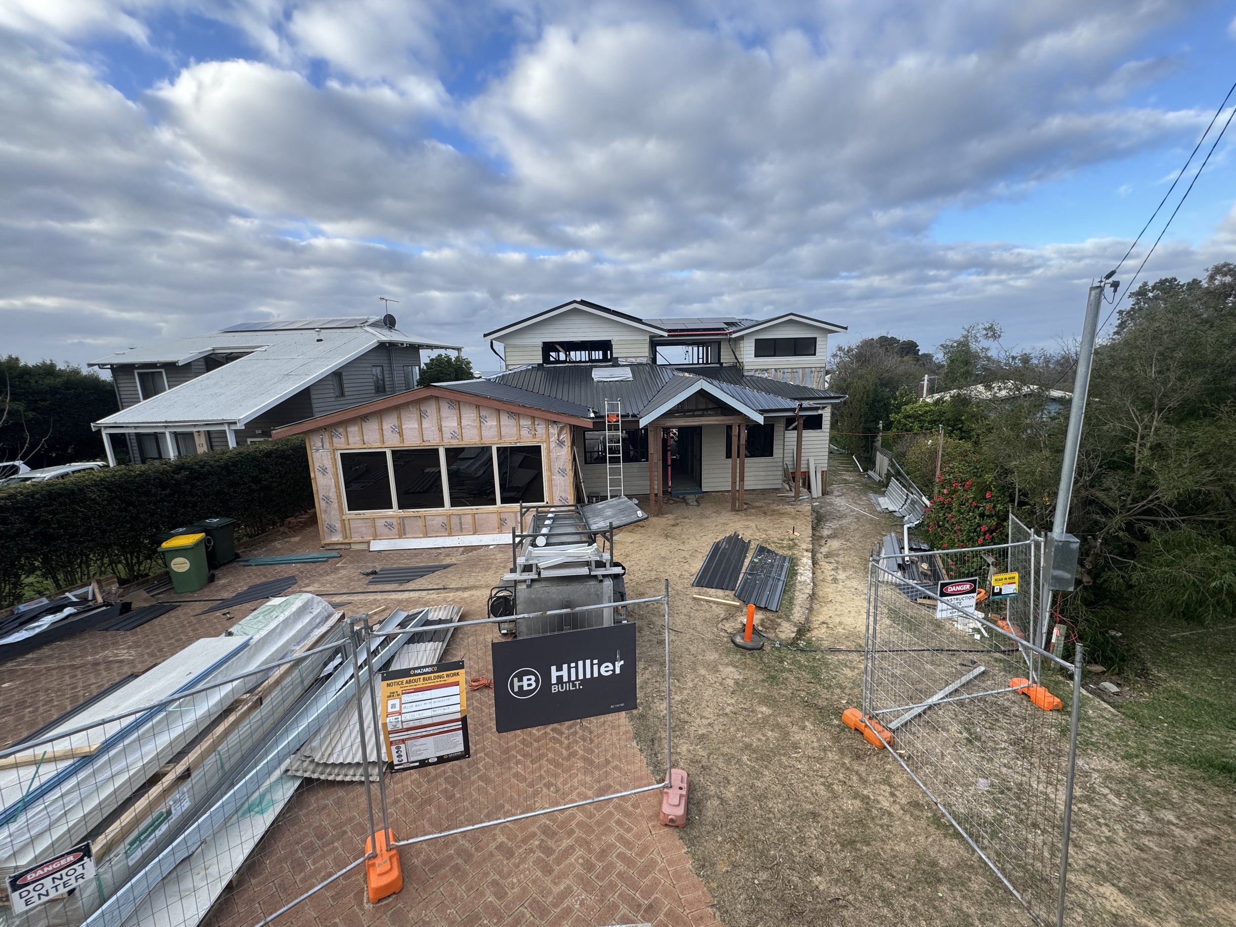 A residential construction site with partially built house structures. The scene includes house frames, construction materials, and safety signs, with cloudy skies overhead.