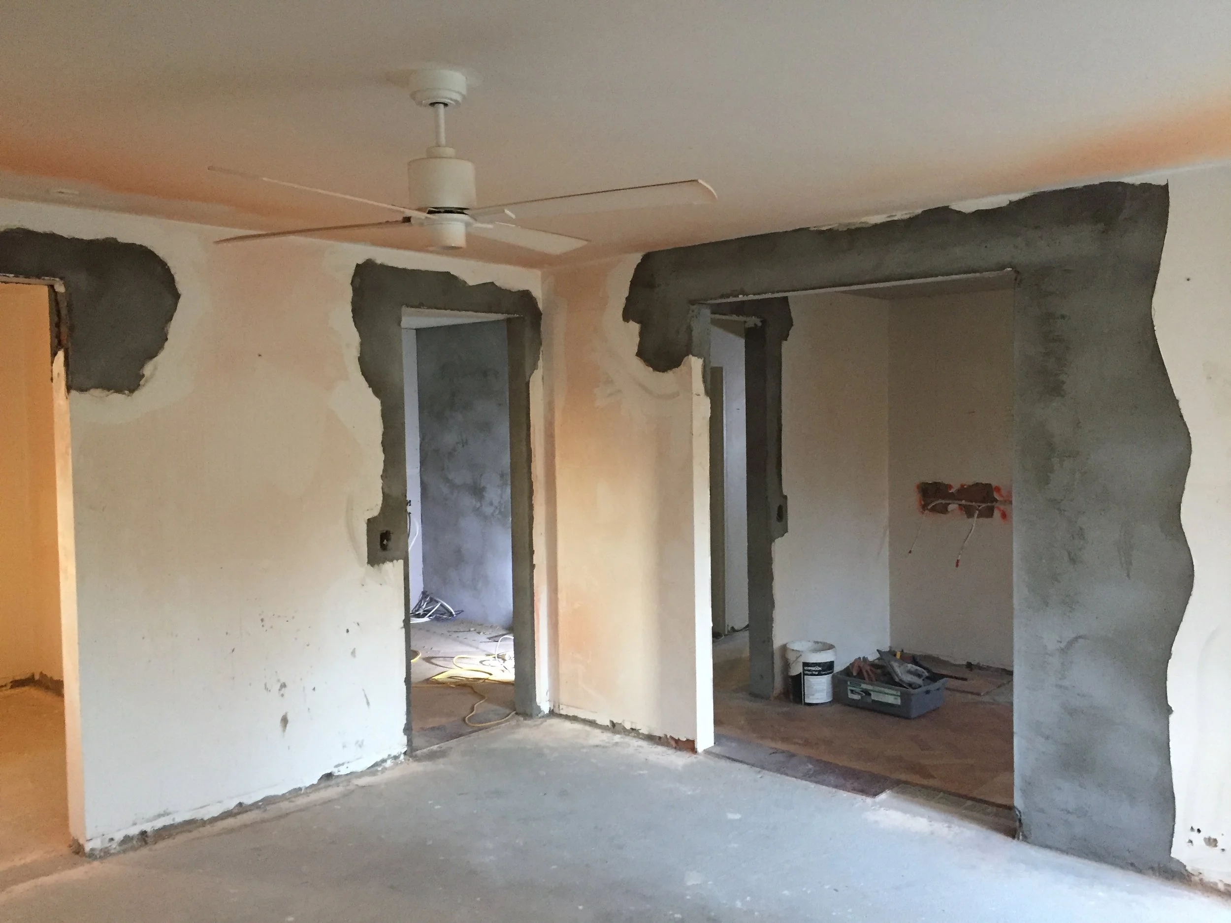 Interior of a room under renovation, with walls partially demolished, construction tools and supplies on the floor, and a ceiling fan installed.