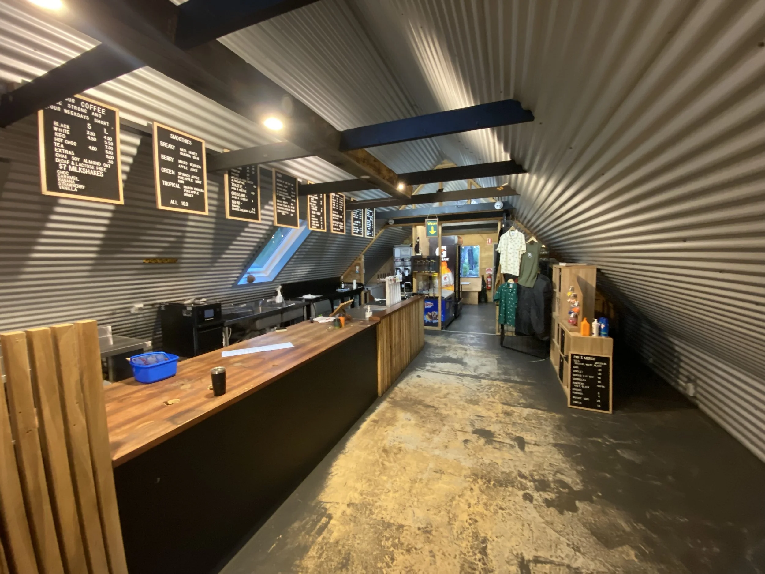 A cafe or coffee shop with corrugated metal walls, wooden accents, a menu on hanging blackboards, a counter with coffee equipment, and casual clothing items displayed.