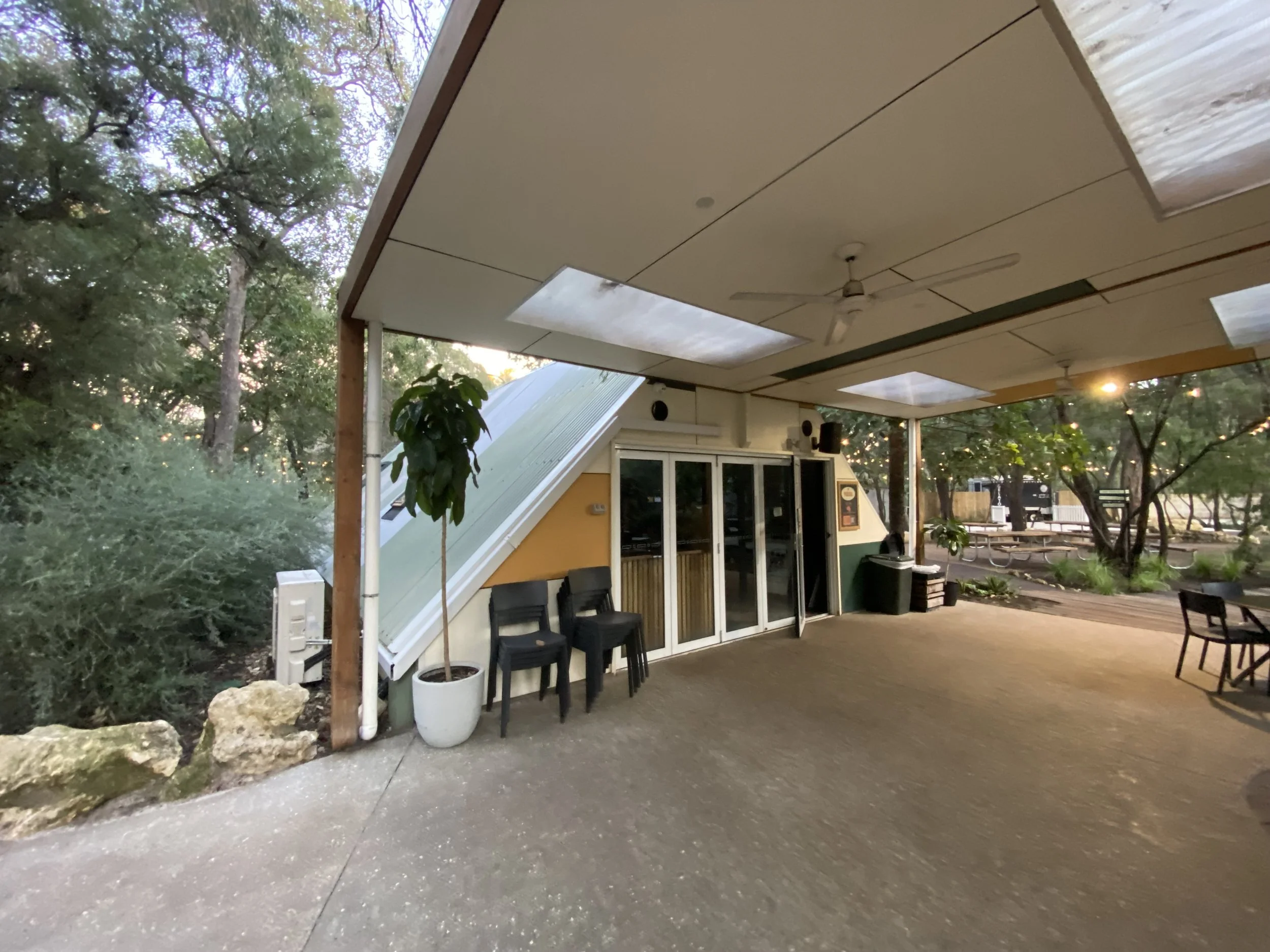 Outdoor patio area with a ceiling fan, chairs, and a yellow building with glass doors, surrounded by trees and greenery.