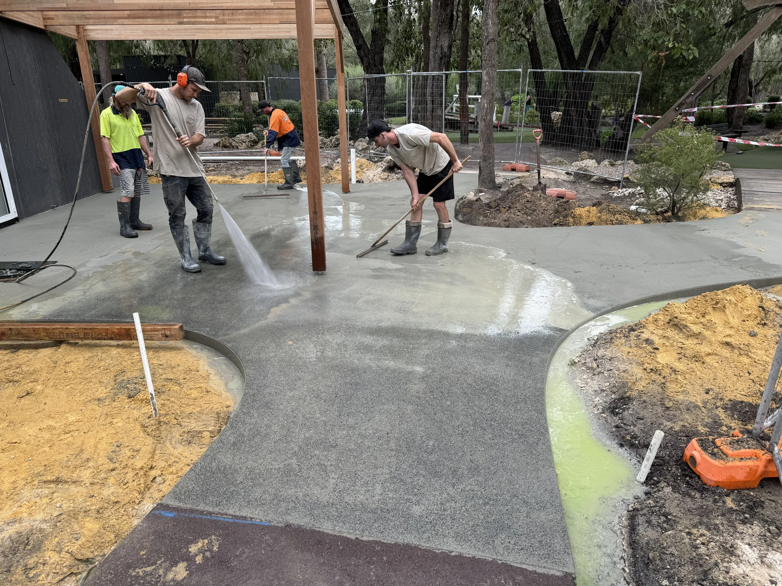 Workers with rubber boots leveling concrete pavement in a construction site surrounded by trees and fencing.