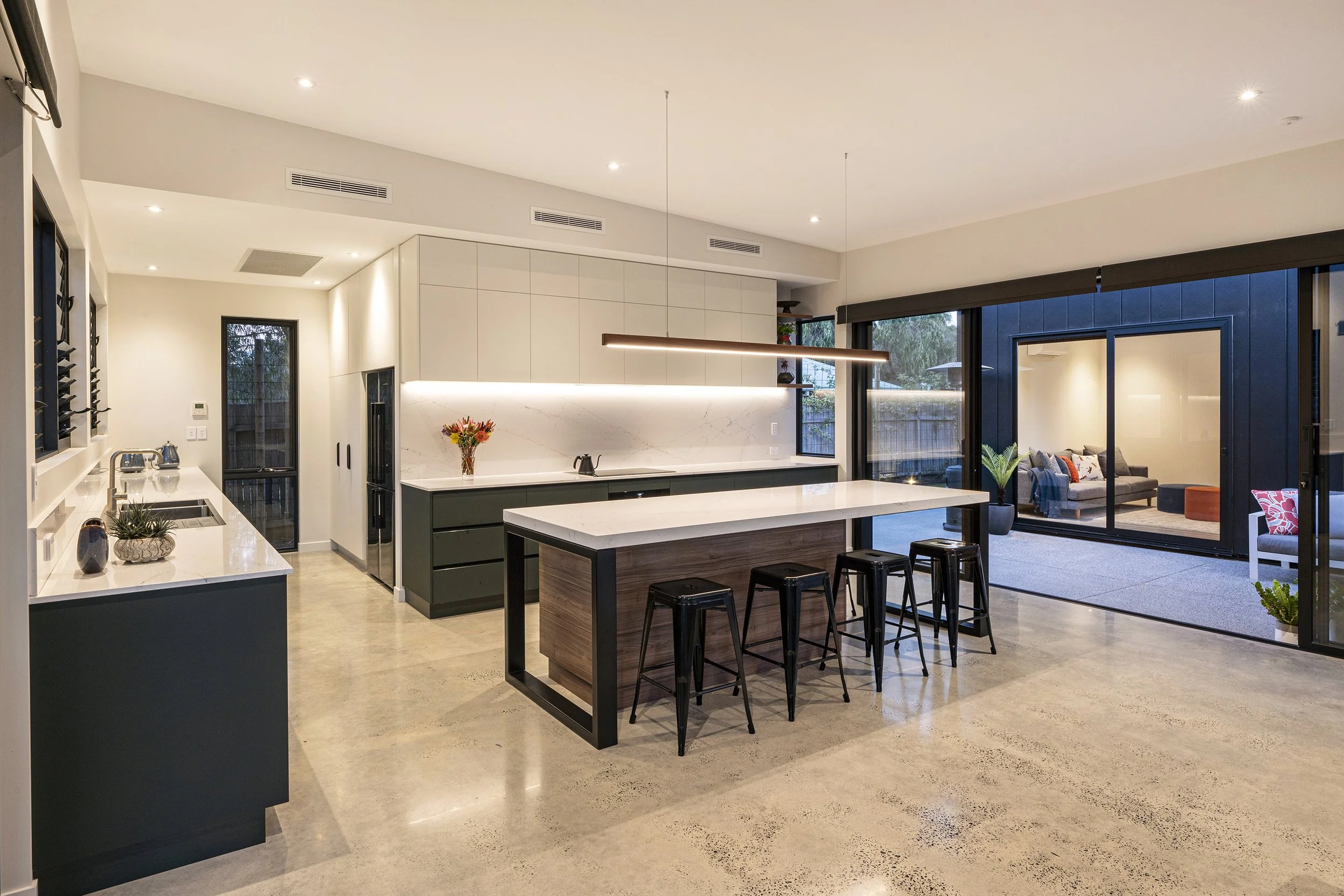 Modern kitchen with white countertops, black cabinets, and a dining area with black stools. Sliding glass doors lead to an outdoor patio with a seating area.