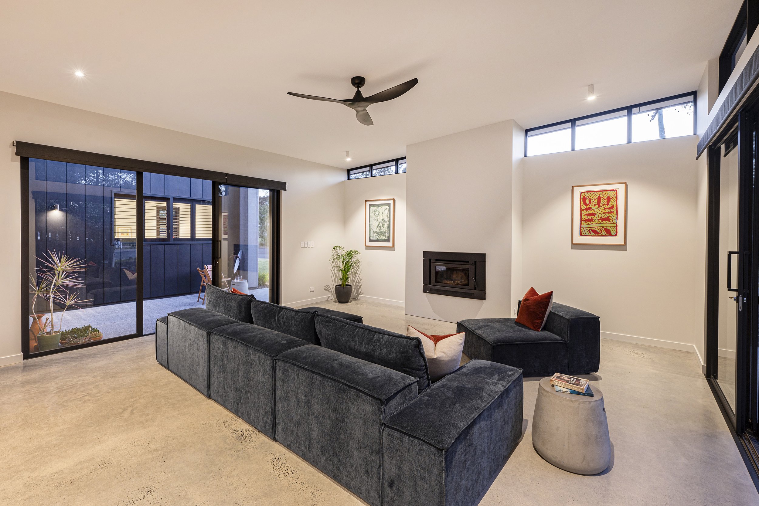 Modern living room with large gray sectional sofa, artwork on the walls, a fireplace, plants, and large sliding glass doors leading to an outdoor patio.