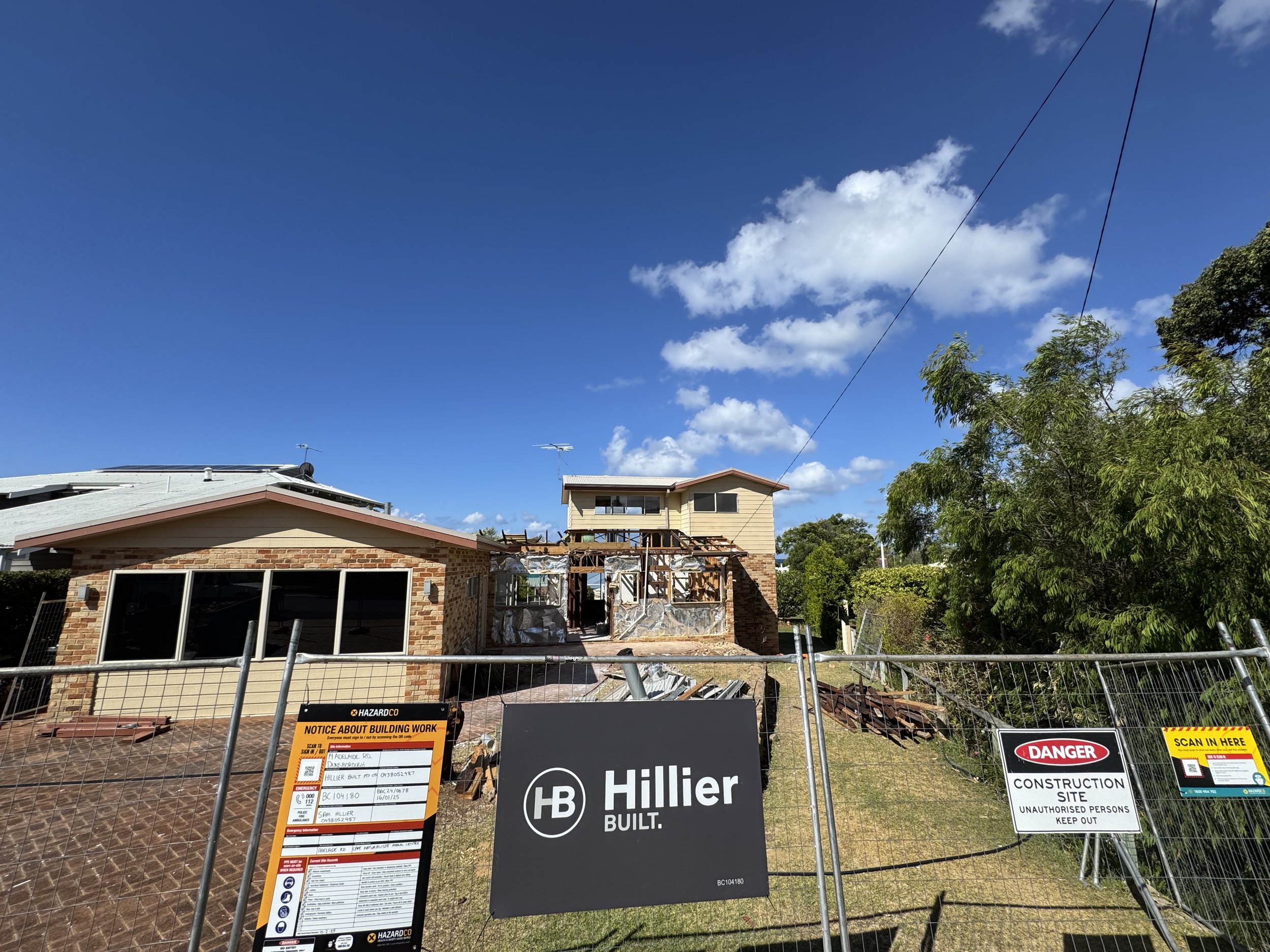 Construction site behind a fence with warning signs, showing a partially built house with exposed framing and a clear blue sky with scattered clouds.