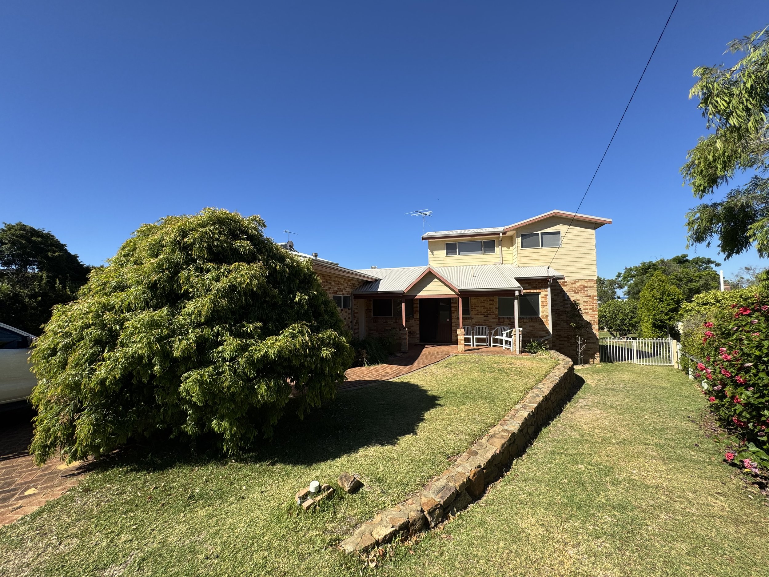 A suburban house with a brick and yellow siding exterior, surrounded by a well-kept lawn and garden with bushes and trees, under a clear blue sky.