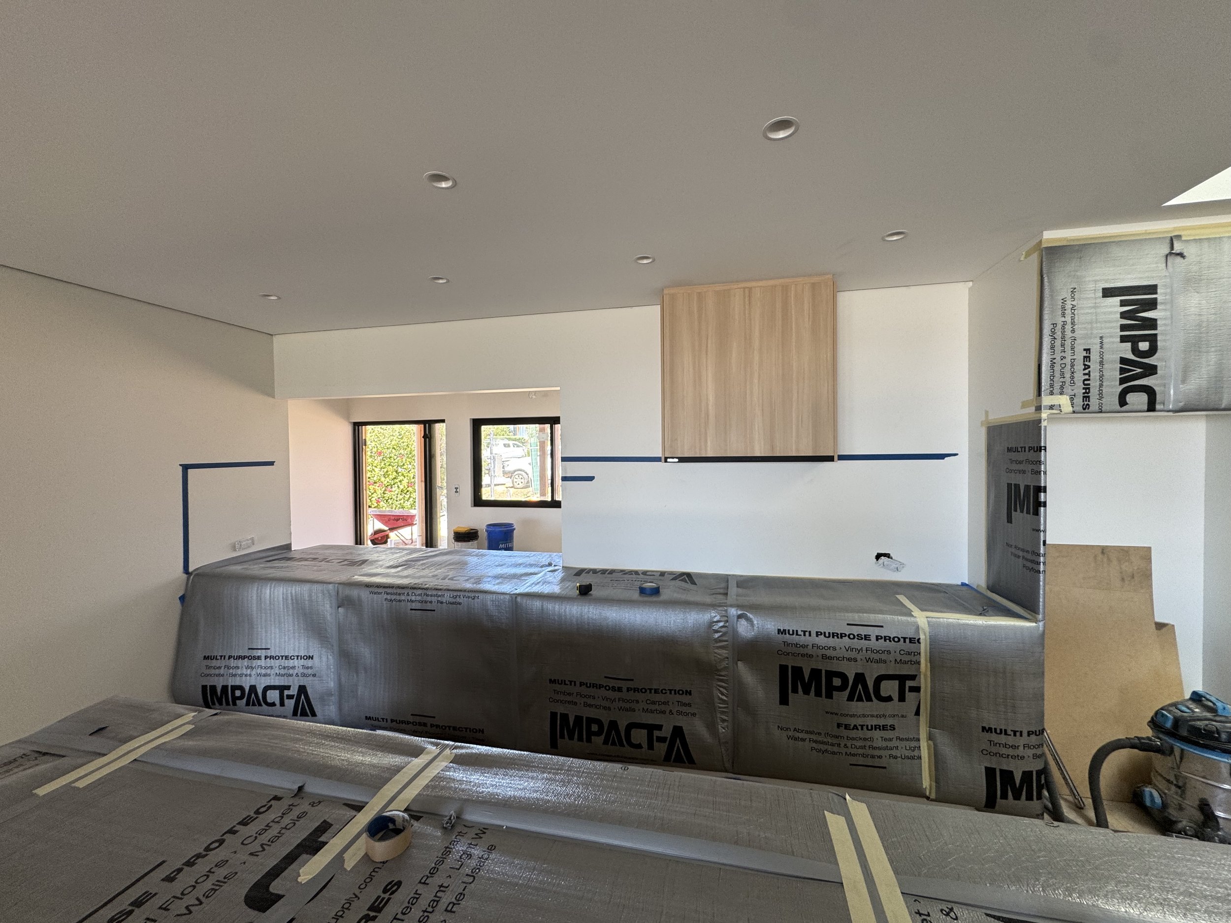 Interior of a room under construction with protective covering on surfaces, several windows allowing natural light, and construction materials and tools visible.