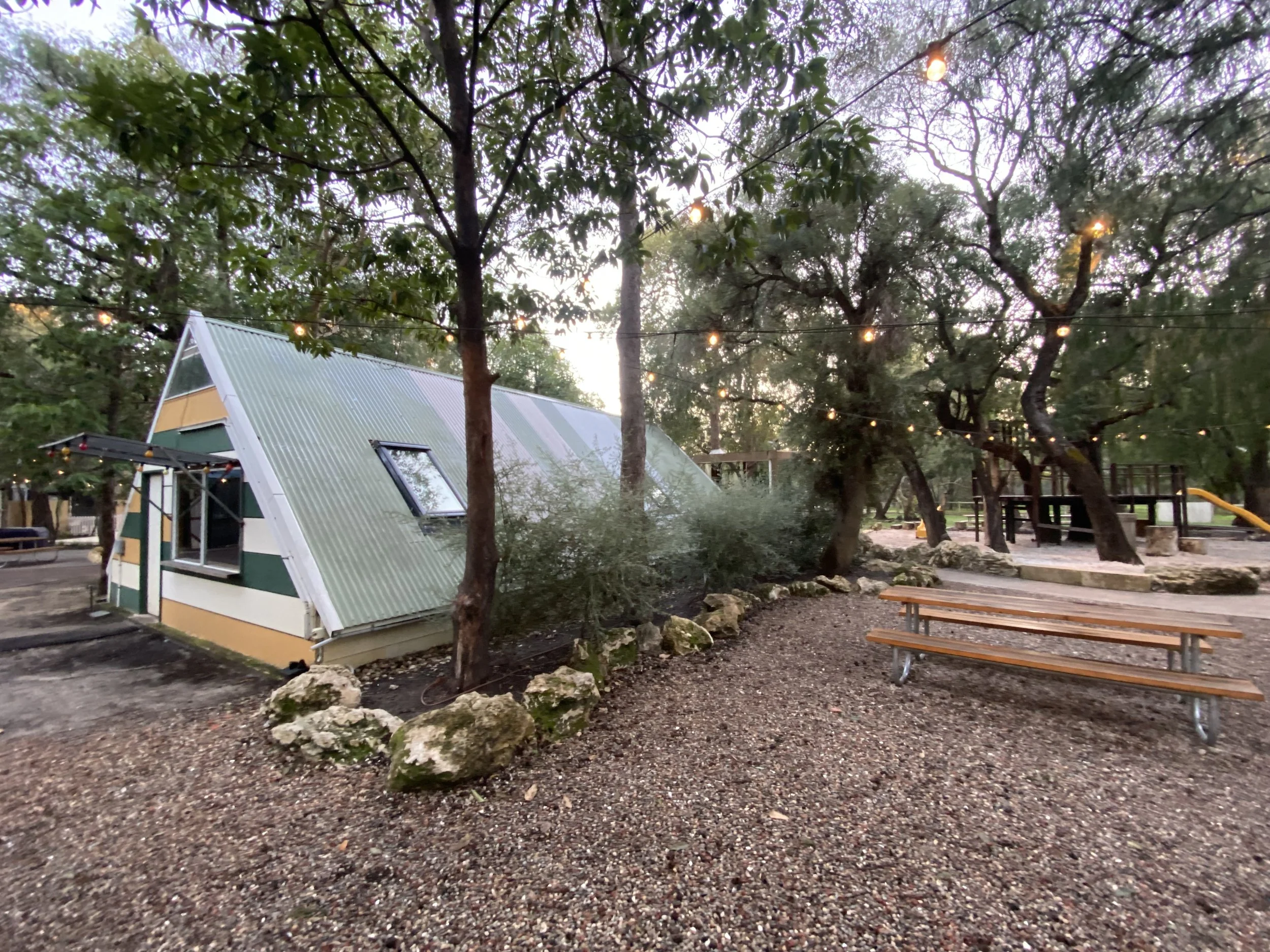 Outdoor scene with trees, string lights hanging between trees, a small building with a slanted roof and window, picnic tables, and ground covered with small rocks.