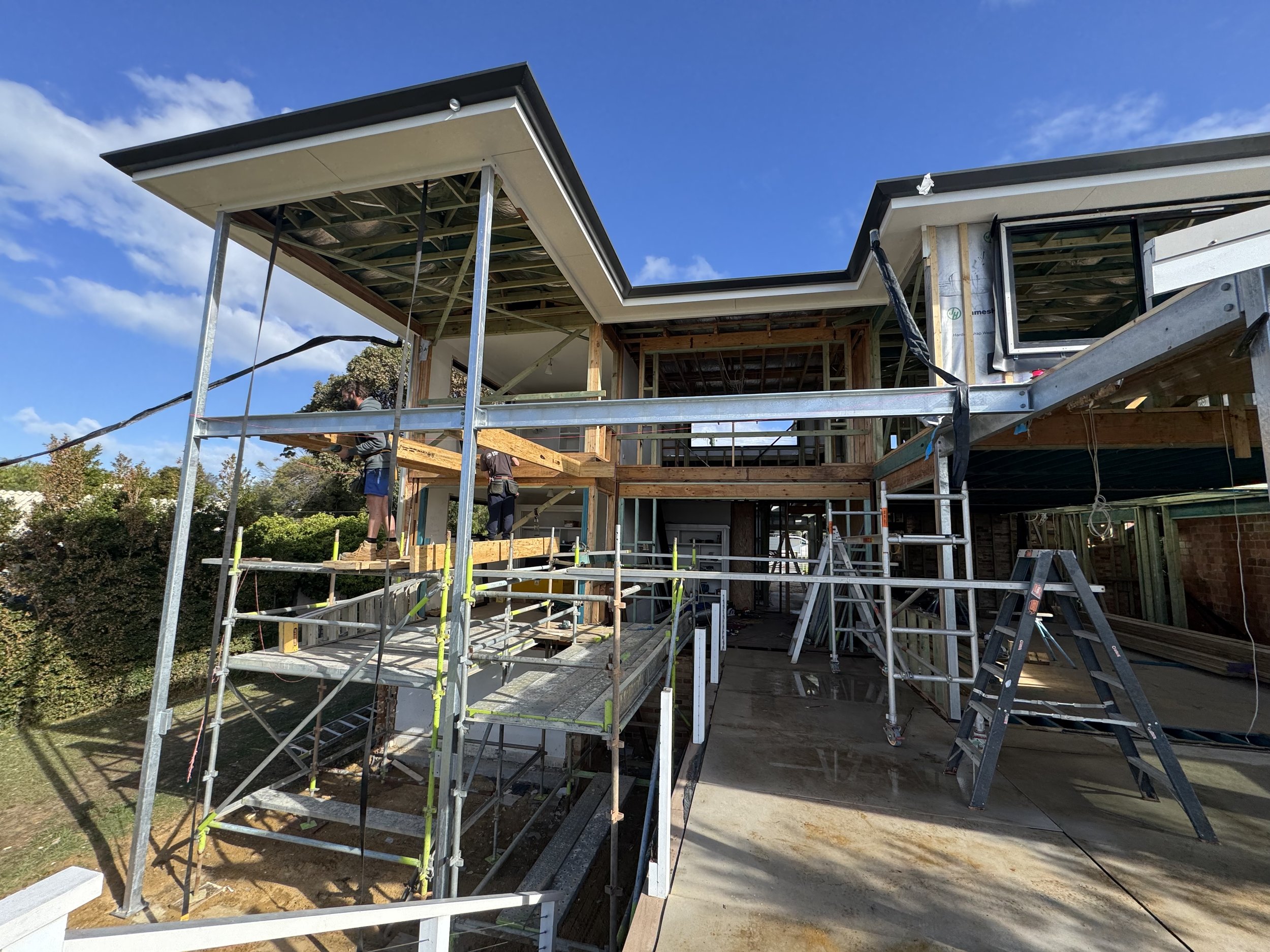 Construction site of a modern house with wood framing, scaffolding, and workers installing siding under a blue sky.