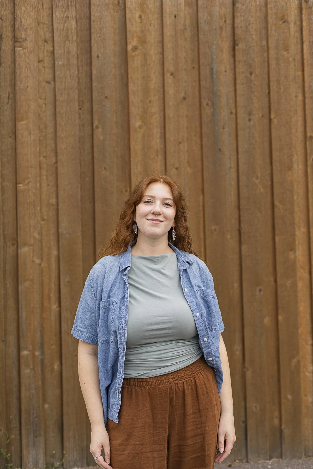 A woman with curly red hair standing in front of a wooden fence, wearing a light green shirt, blue denim shirt, and brown pants.