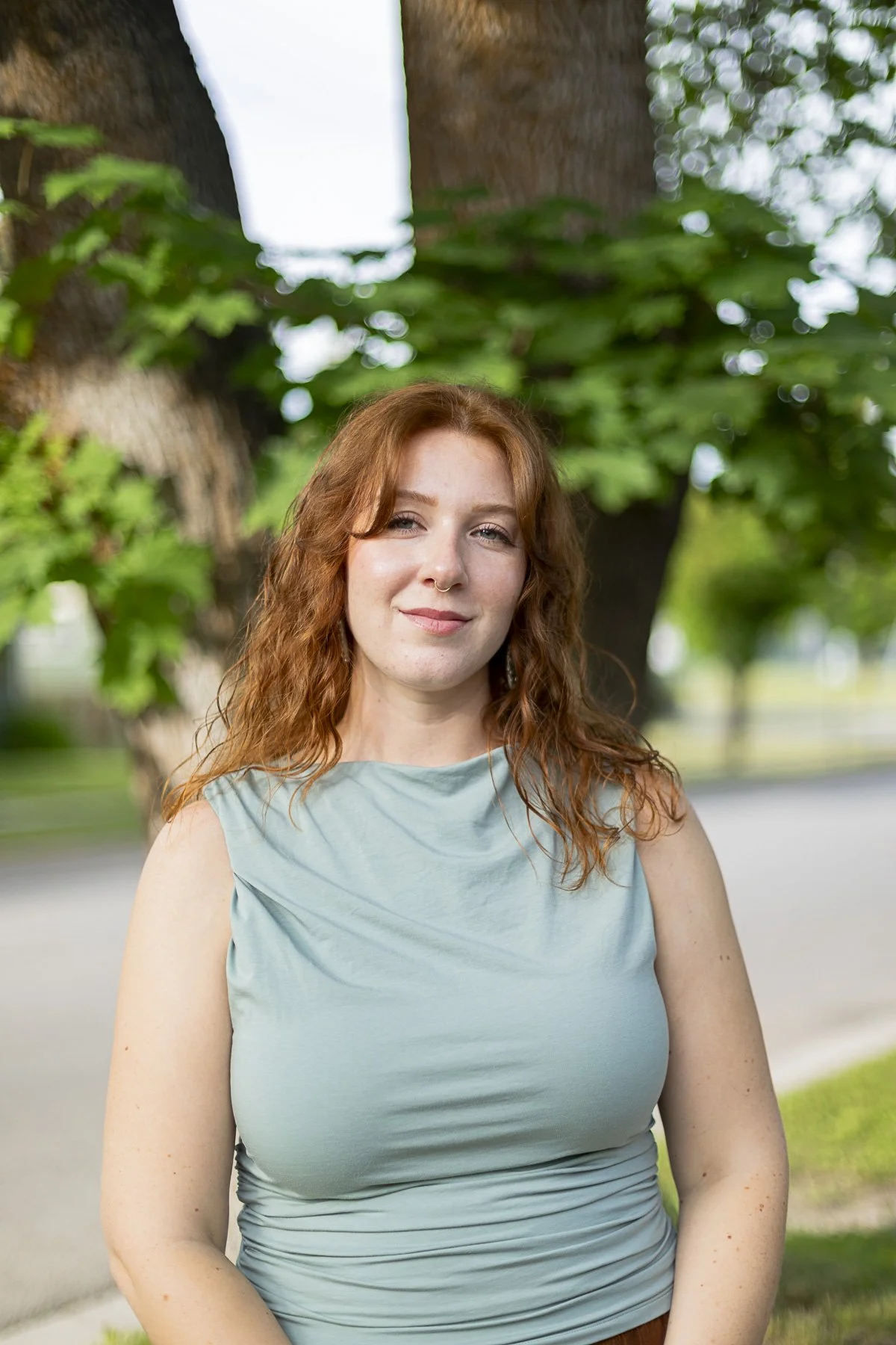 Portrait of a young woman with reddish curly hair and a light blue sleeveless top standing outdoors near a large tree with green leaves.