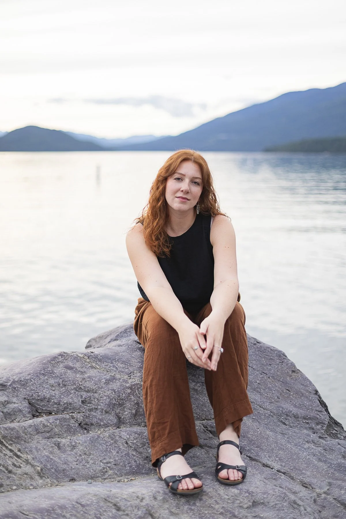 A woman with red hair sitting on a rock by a body of water with mountains in the background, wearing a black sleeveless top, brown pants, and black sandals.