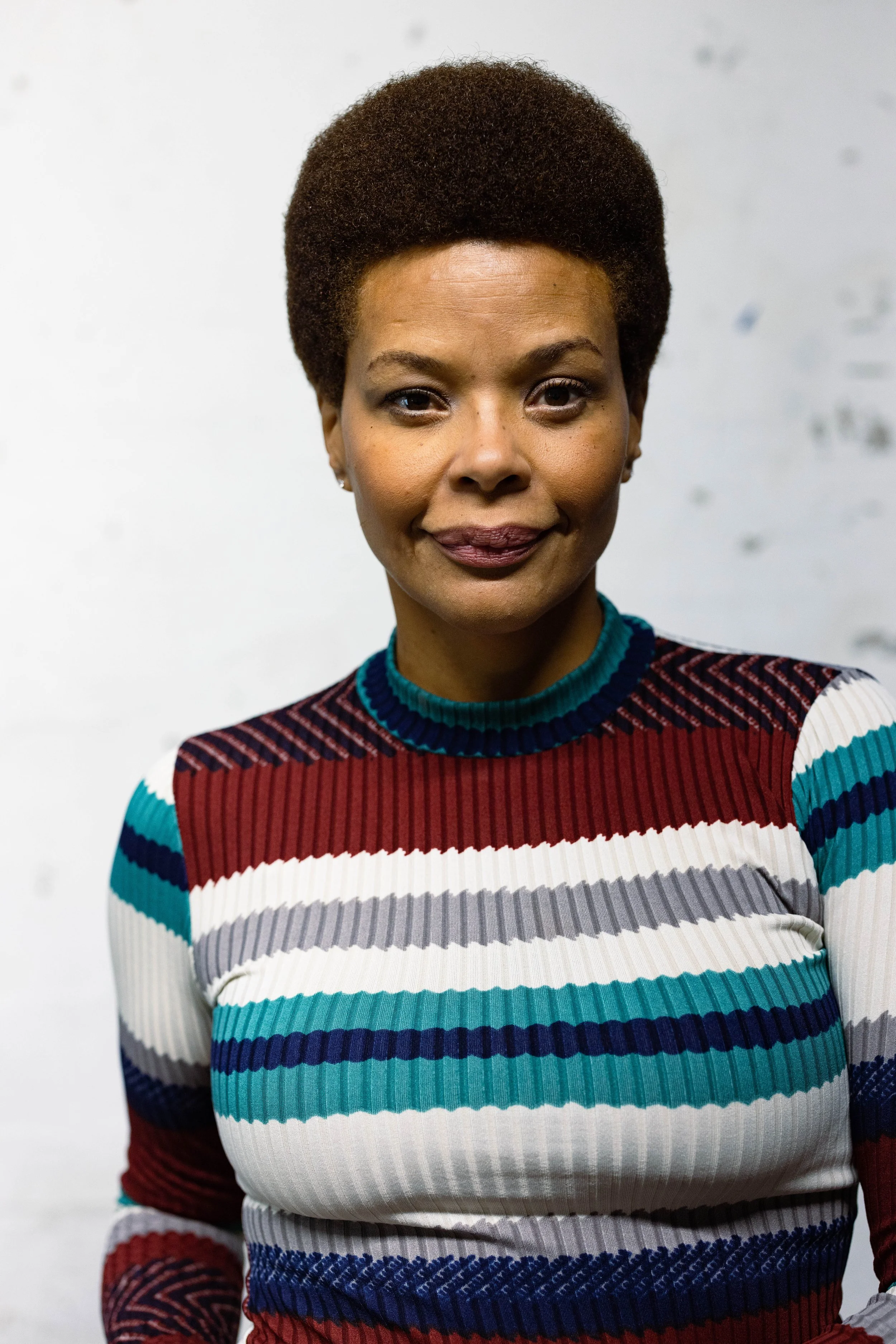 Erickka Jones with natural afro hairstyle wearing a multicolored striped sweater standing in front of a plain white background for an acting headshot. 