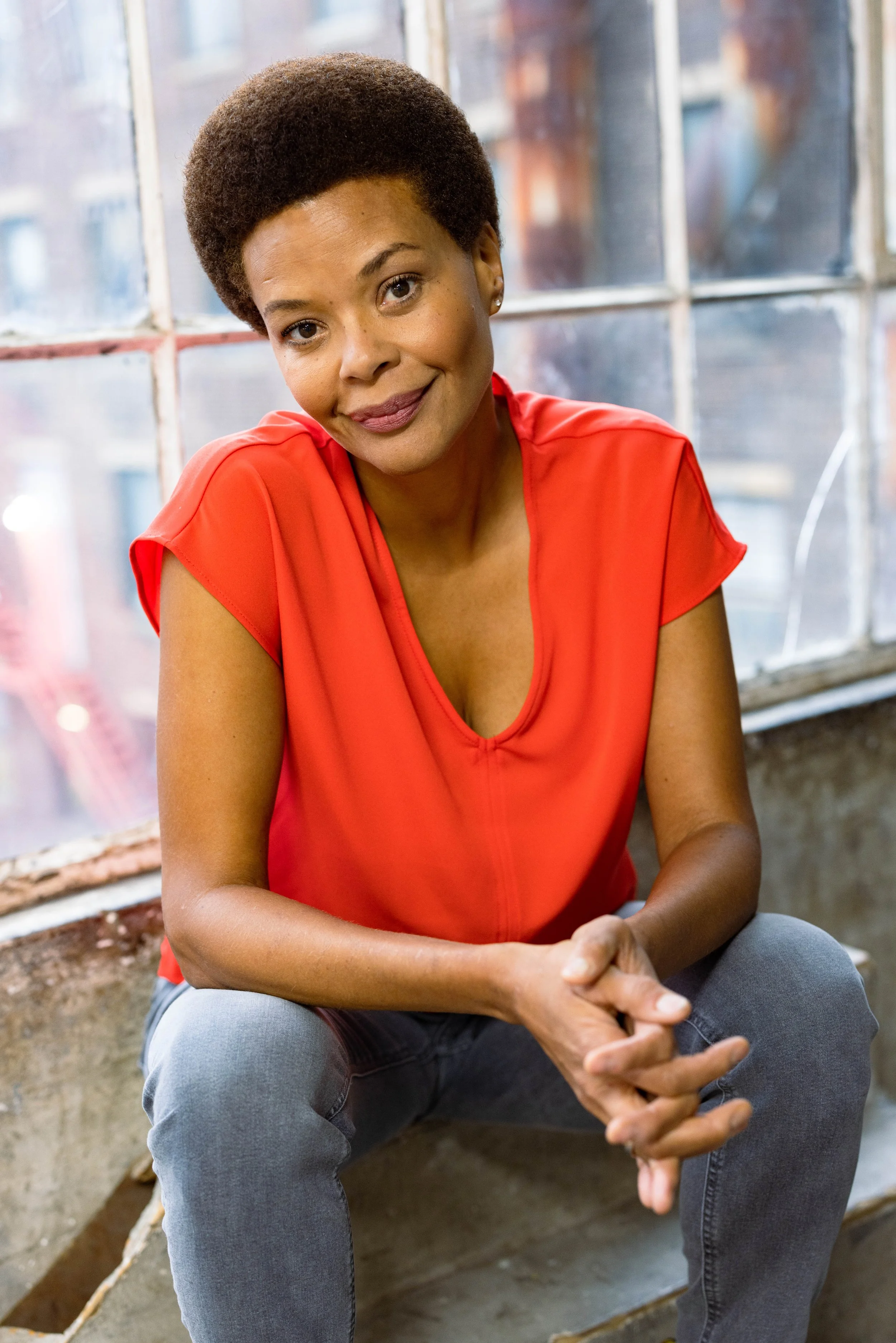 A woman, Erickka Jones, wearing a red top and gray jeans sitting on a concrete step in front of a large window, smiling and looking at the camera.