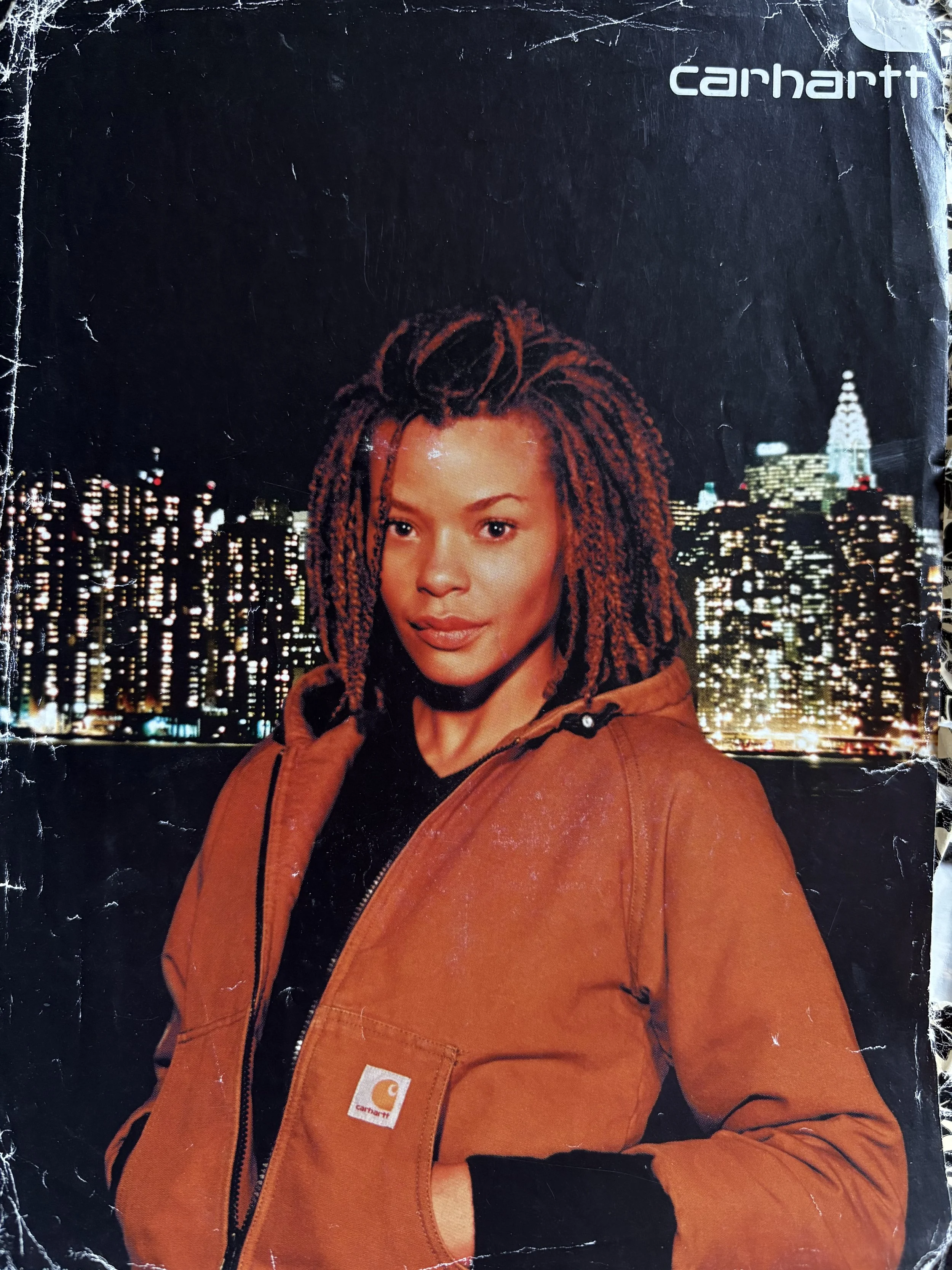 Erickka with locs wearing an orange Carhartt jacket poses in front of a city skyline at night for an international Carhartt ad.