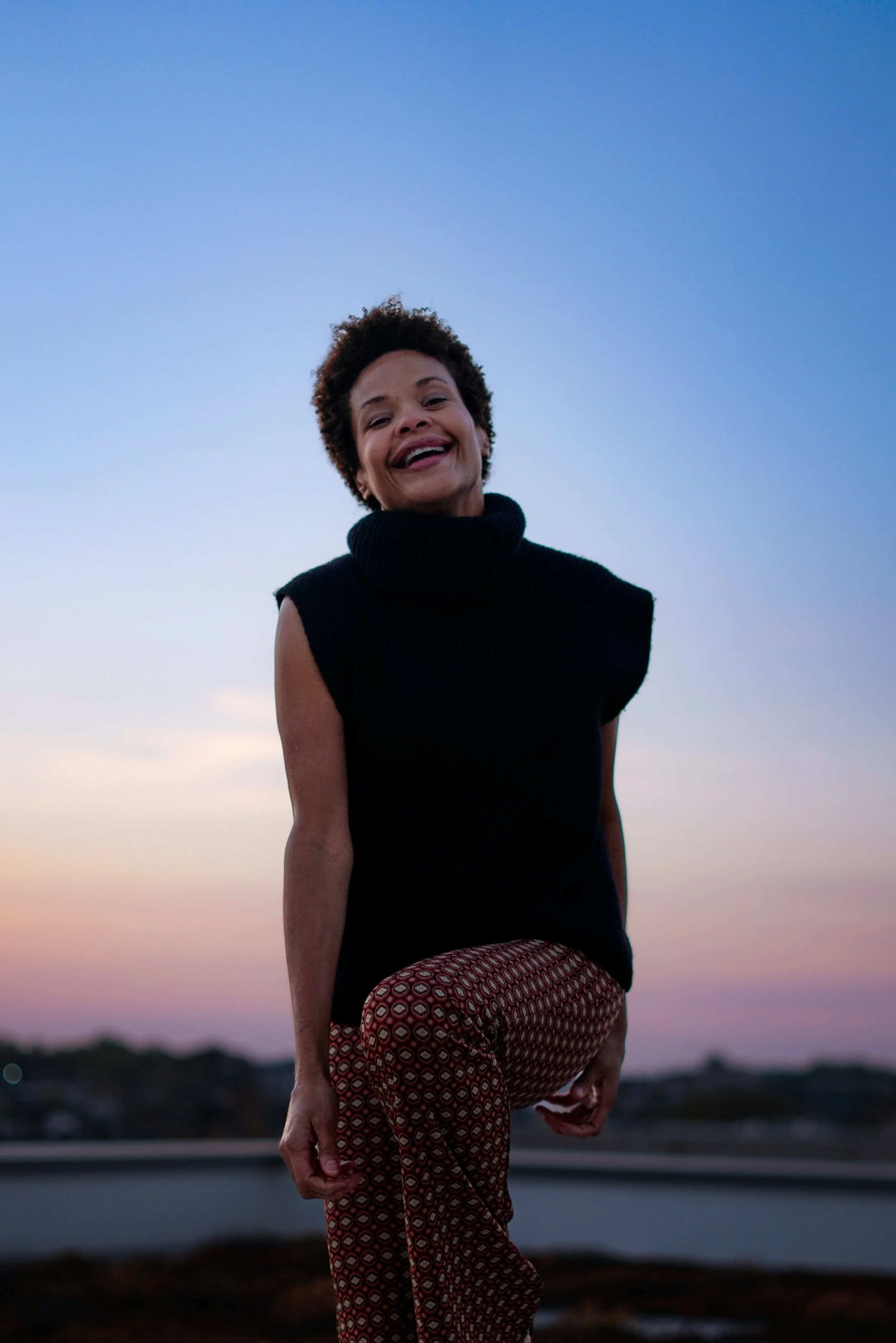 A woman, Erickka Jones, with short curly hair smiling while balancing on her right leg, wearing a sleeveless black turtleneck top and patterned pants, outdoors during sunset or dusk with a blue sky and distant landscape.