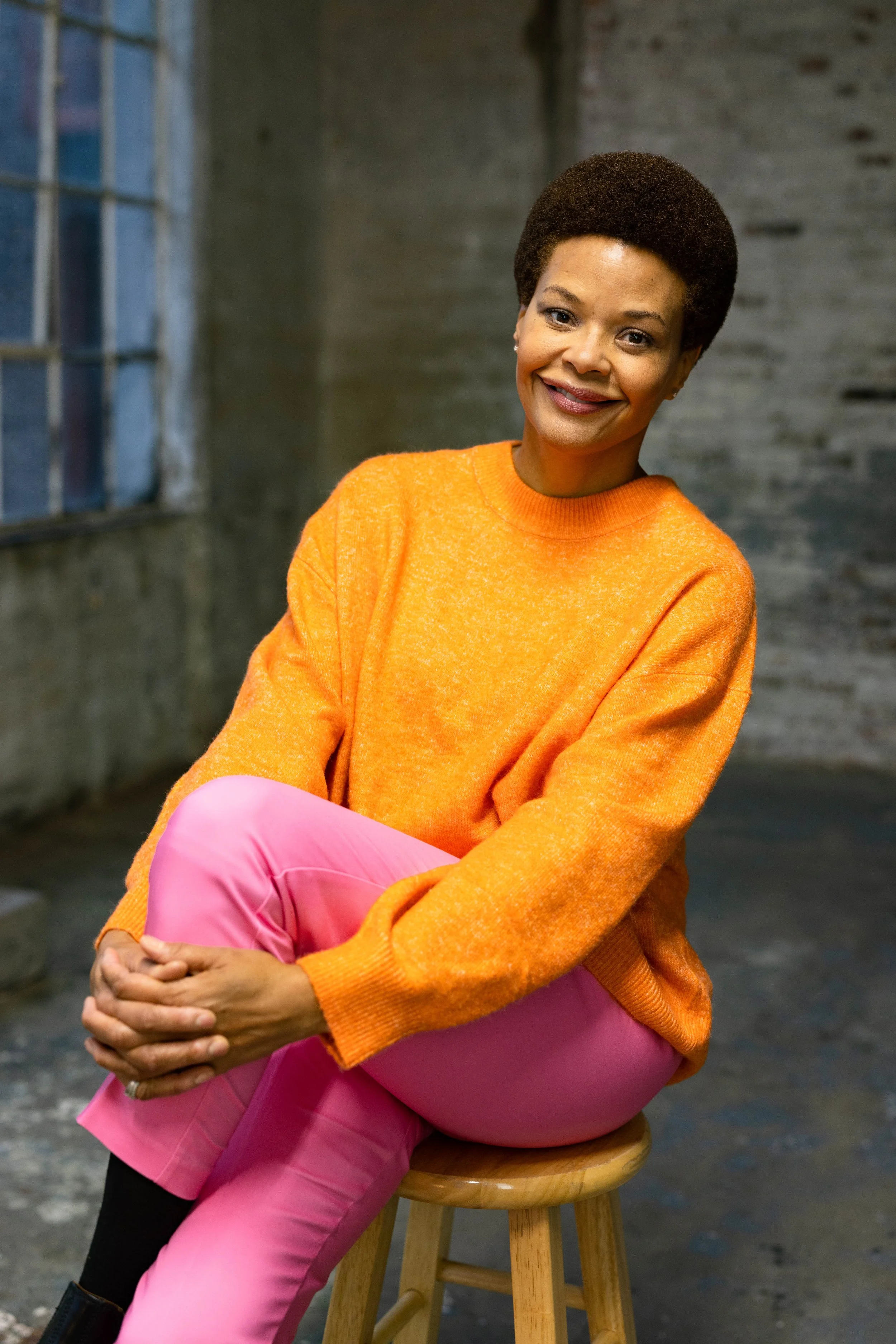 A smiling woman with short curly hair, wearing an orange sweater and pink pants, sitting on a wooden stool against a gray brick wall and window background.