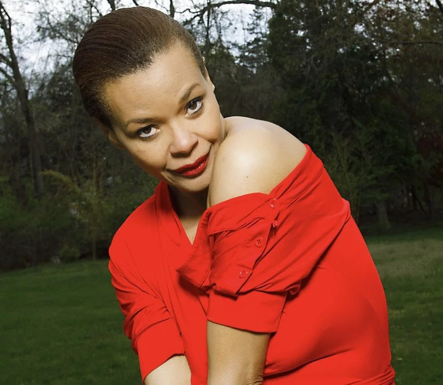 A woman with short hair and red lipstick, wearing a red off-shoulder top, standing outdoors in a grassy area with trees in the background.
