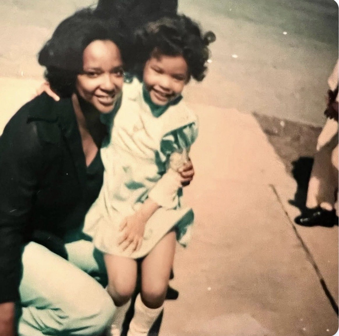 A mom (Sarah Jones) and daughter (Erickka Jones) curly hair smiling, one wearing a black jacket and the other in a white coat, sitting or crouching outdoors on a sidewalk.