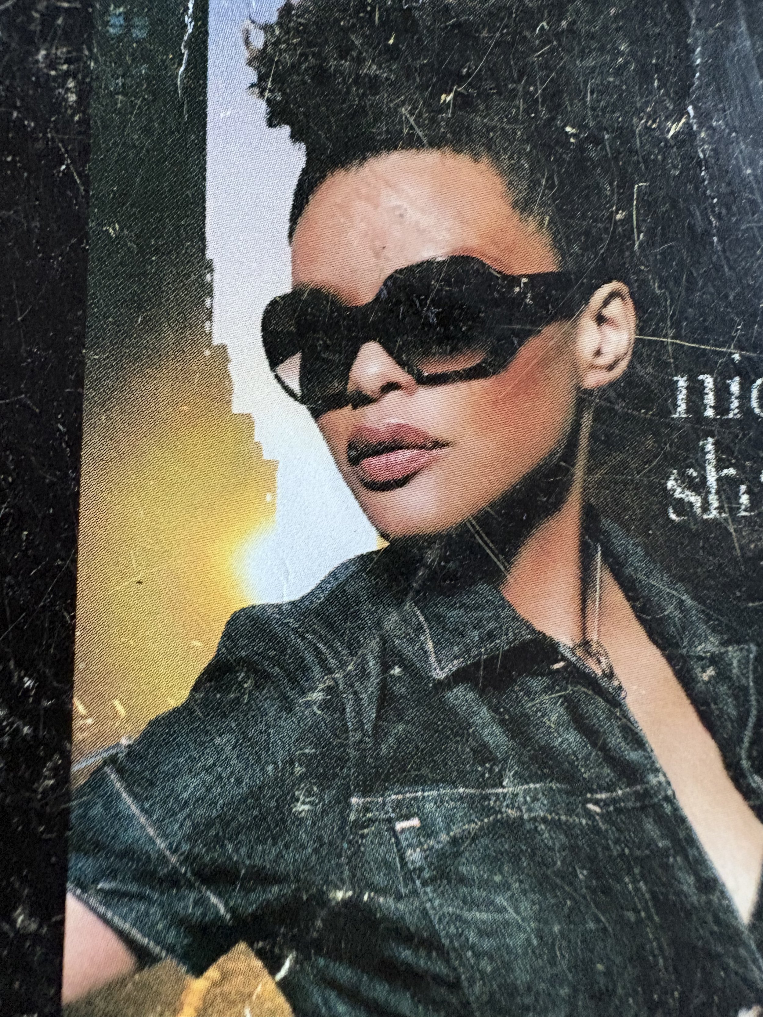 A woman wearing sunglasses with a patterned shirt, dark hair styled up, with a background that includes a gradient of blue and yellow.